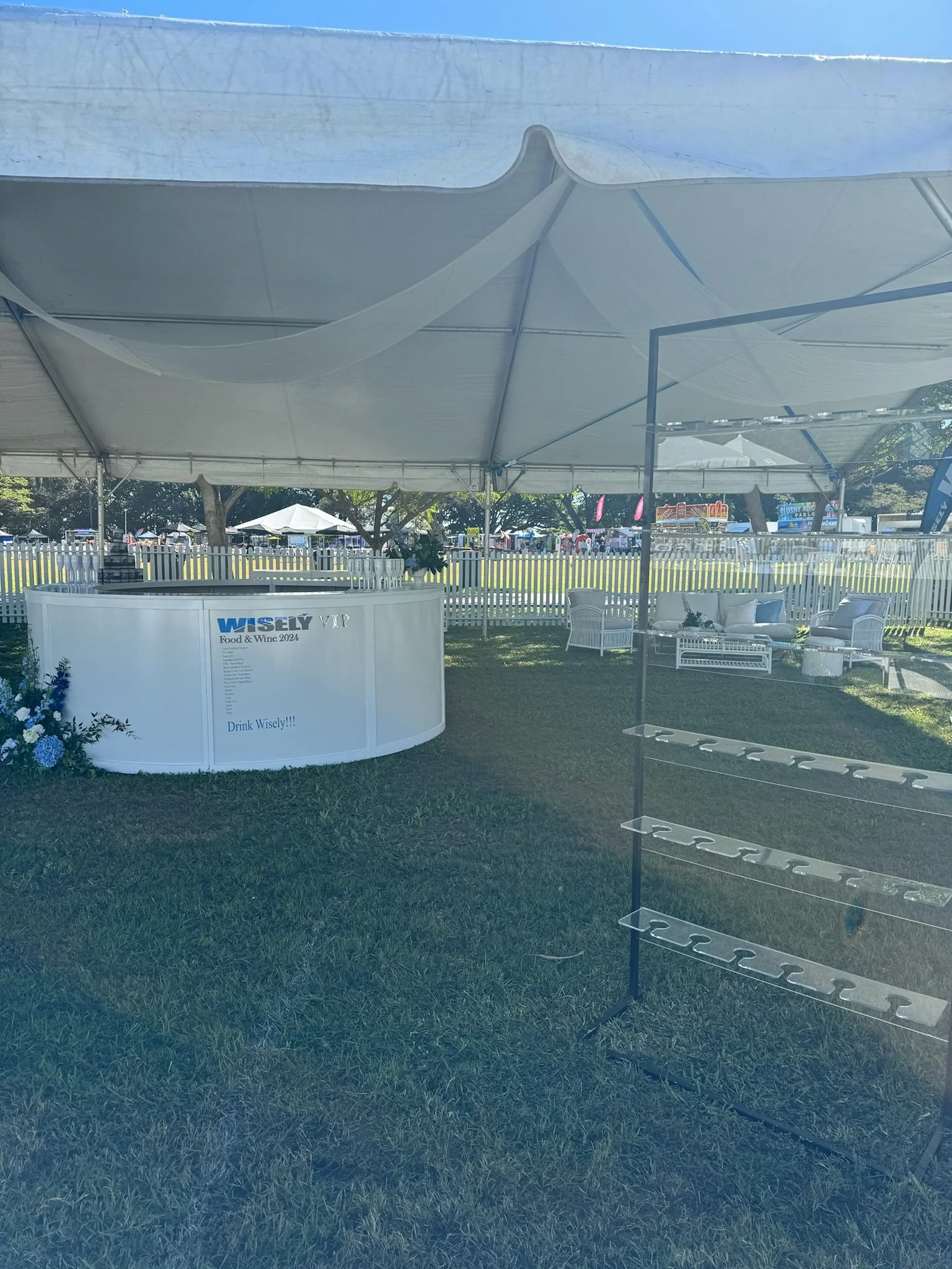 Empty outdoor event tent with white fabric, some chairs and a display on a grassy field at a festival or fair, with tents and people in the background.