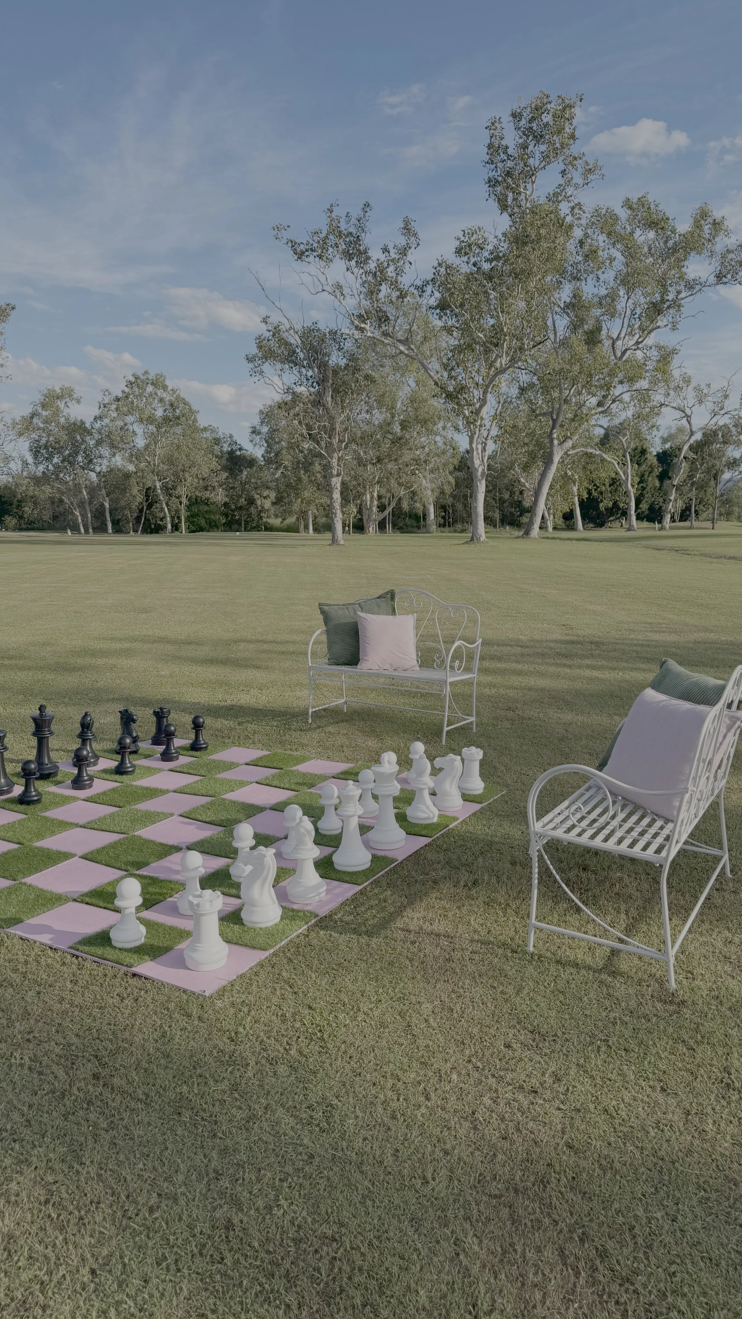 A large open grassy field with scattered trees under a blue sky with some clouds. There are two white chairs with cushions and a decorative chessboard with large chess pieces on the grass.