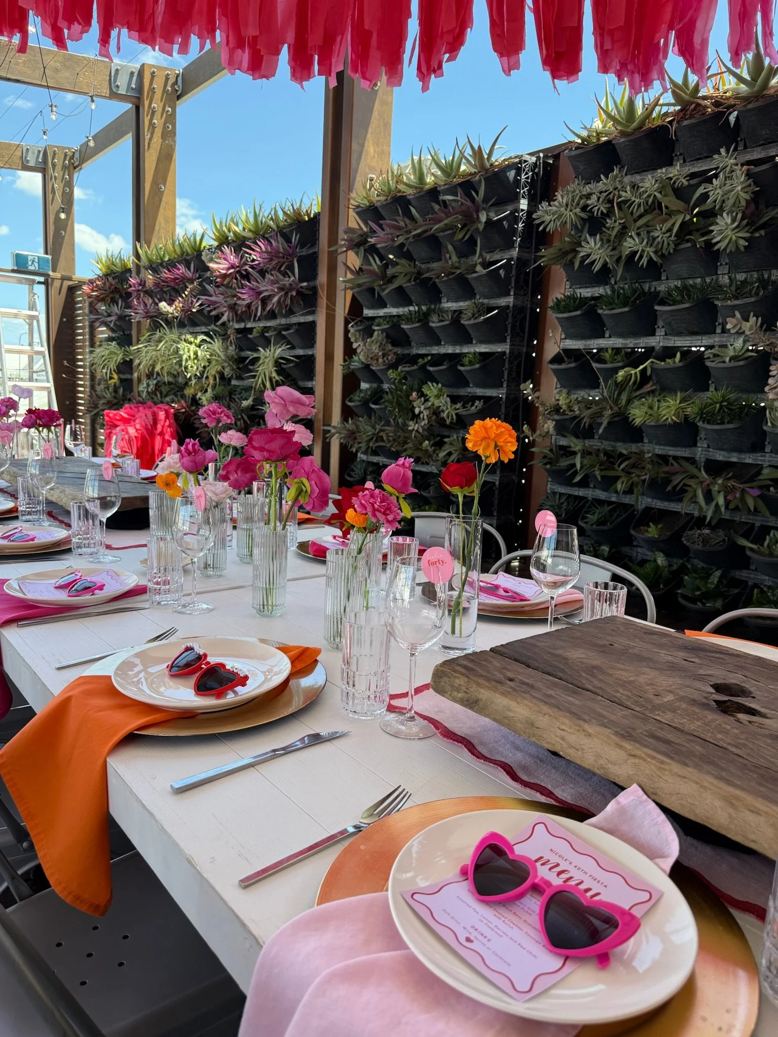 A decorated outdoor table set for a celebration, with colorful flowers in vases, pink and orange sunglasses, and place settings including plates, napkins, glasses, and cutlery, under a pink and red paper canopy, with a green wall of succulents in the