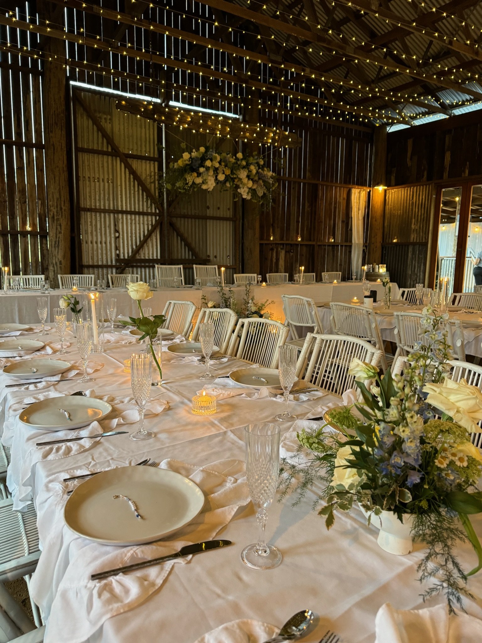 A decorated rustic wedding reception with a long table set with white plates, crystal glasses, floral centerpieces, and candles. The setting is inside a barn with wooden walls and exposed beams, adorned with fairy lights and a hanging flower arrangem