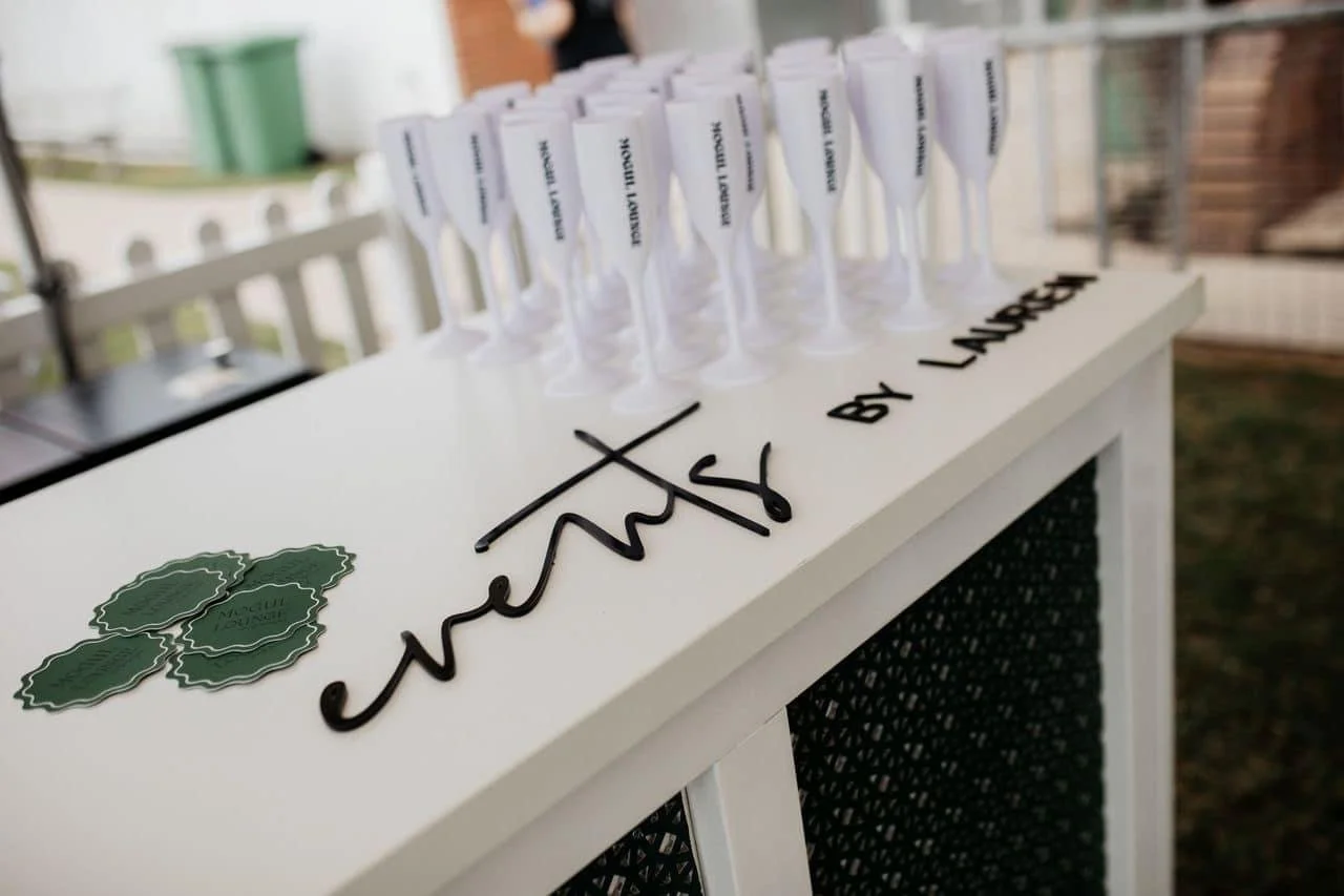 A white table with mini champagne bottles and black text that says "Congratulations by Lola". The table is outdoors with a blurred background.