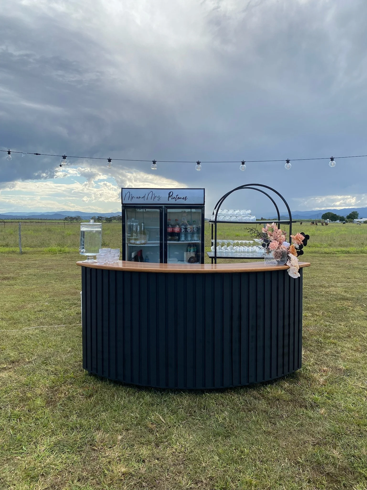 A black outdoor drink cart with a wooden top, decorated with pink and white flowers, standing in a grassy field under a cloudy sky. There is a glass water dispenser and cups on the cart, with a small refrigerator in the background. String lights hang