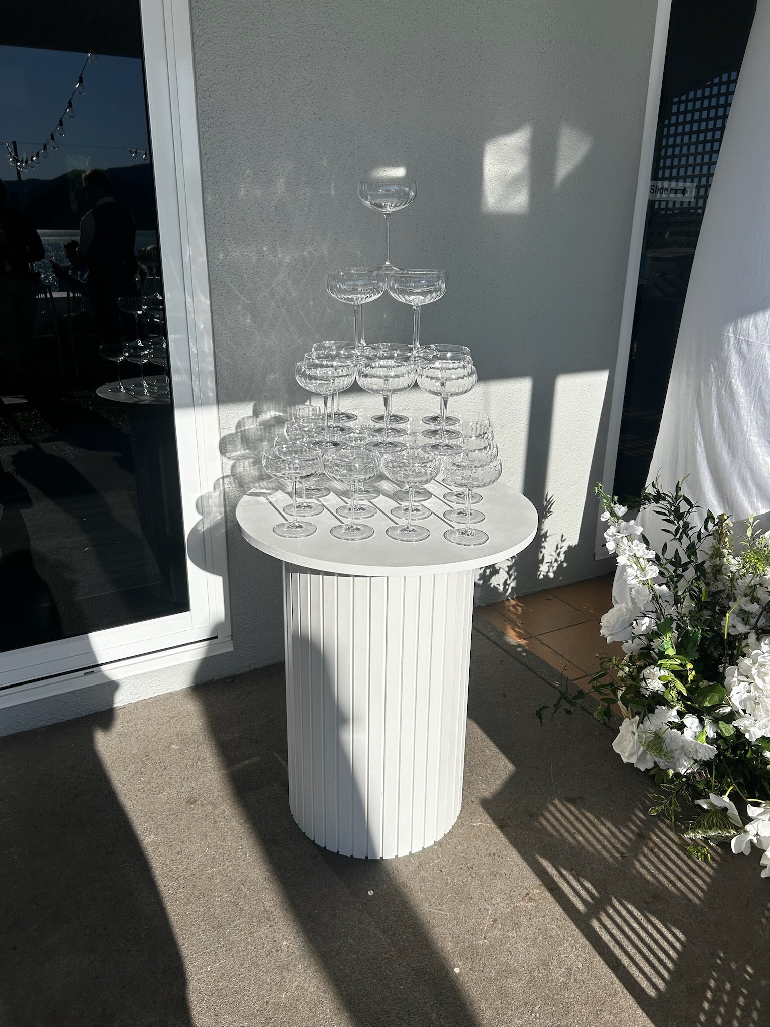 Stacked champagne glasses arranged in a pyramid on a white cylindrical pedestal outside, with sunlight and shadows.