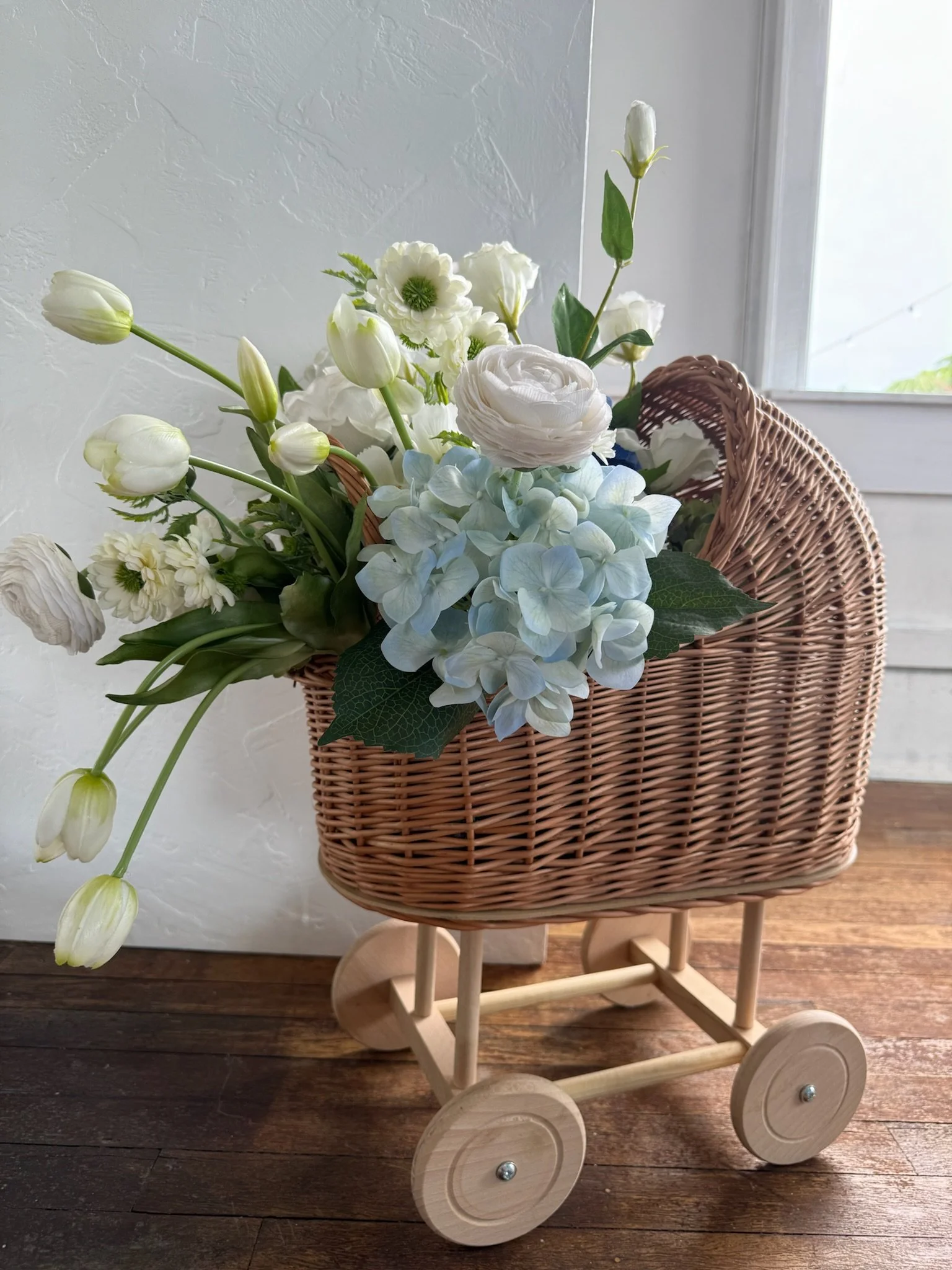 A wicker basket filled with white and light blue flowers sits on a wooden doll stroller near a window.