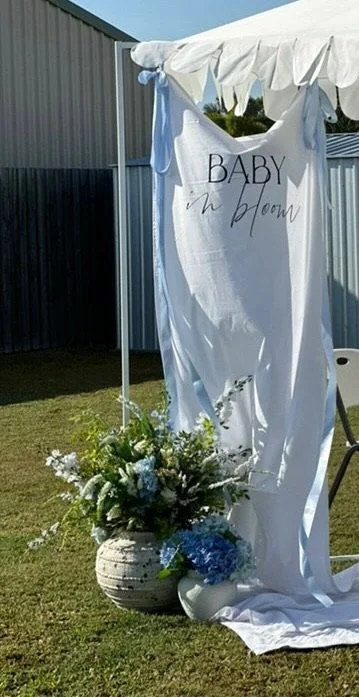 Outdoor baby shower setup with a white fabric backdrop, a floral arrangement in a large ceramic vase, and a sign that reads 'Baby in Bloom'.