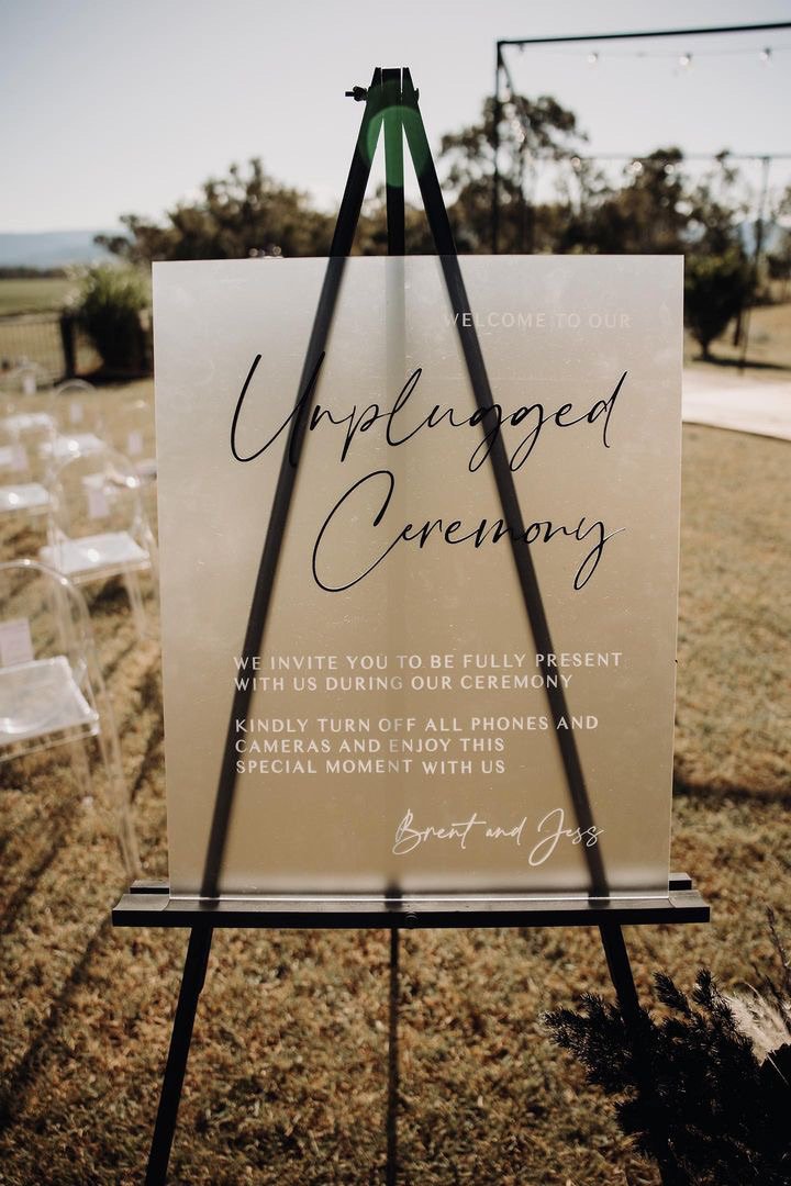 Sign on a stand reading 'Unplugged Ceremony,' inviting guests to be fully present and turn off phones during the wedding of Brent and Jess, with an outdoor setting in the background.