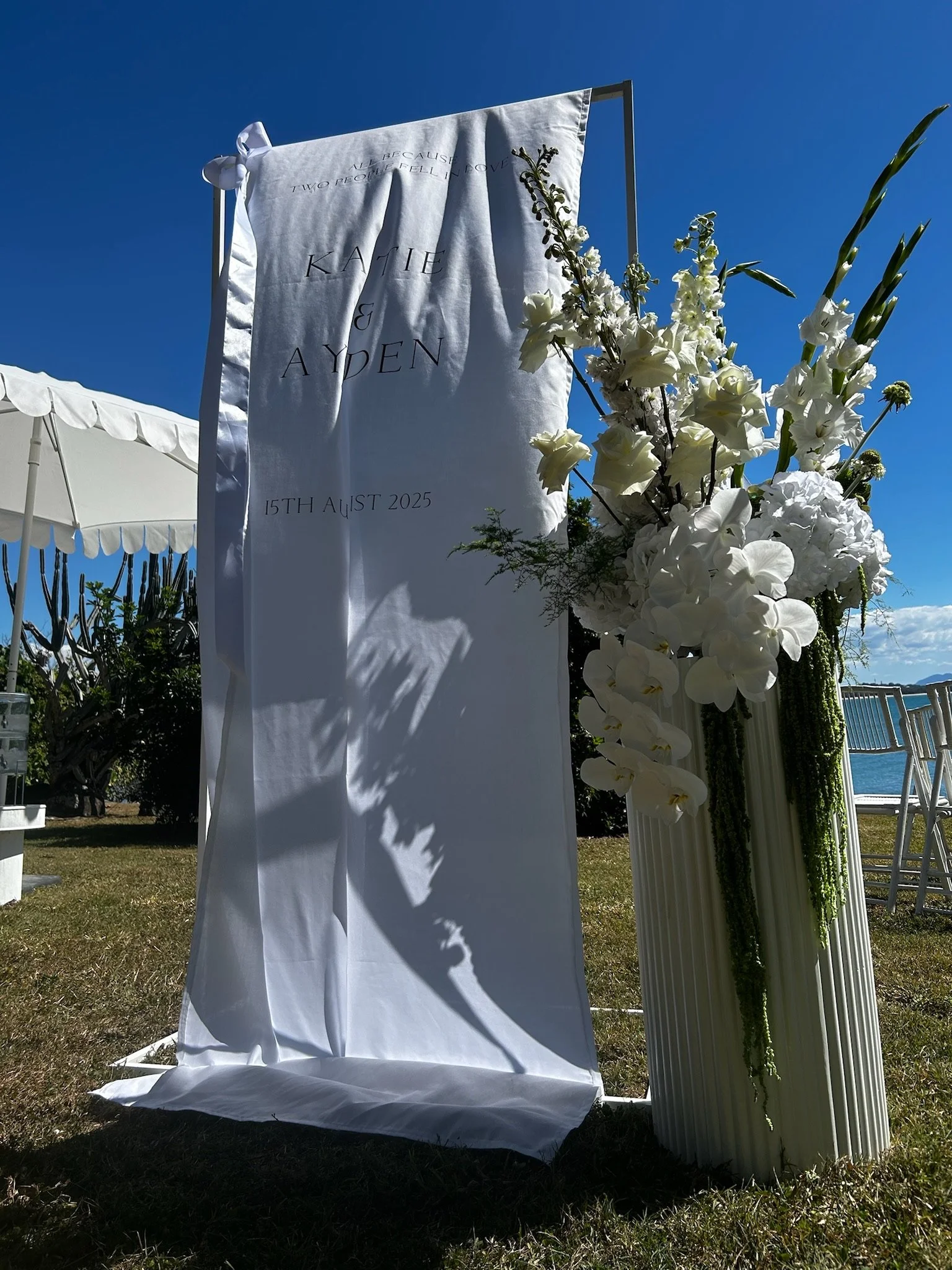 White wedding banner with names Katie and Aydon, and wedding date August 15, 2025, next to a tall floral arrangement of white flowers on a striped white pedestal, outdoors under a clear blue sky.