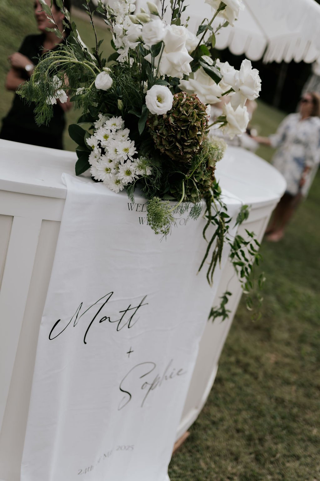 Close-up of a white wooden welcome table with a floral arrangement of white and green flowers and greenery, and a fabric banner with handwritten-style text that reads 'Matt + Sophie'. In the background, two women are talking outdoors.