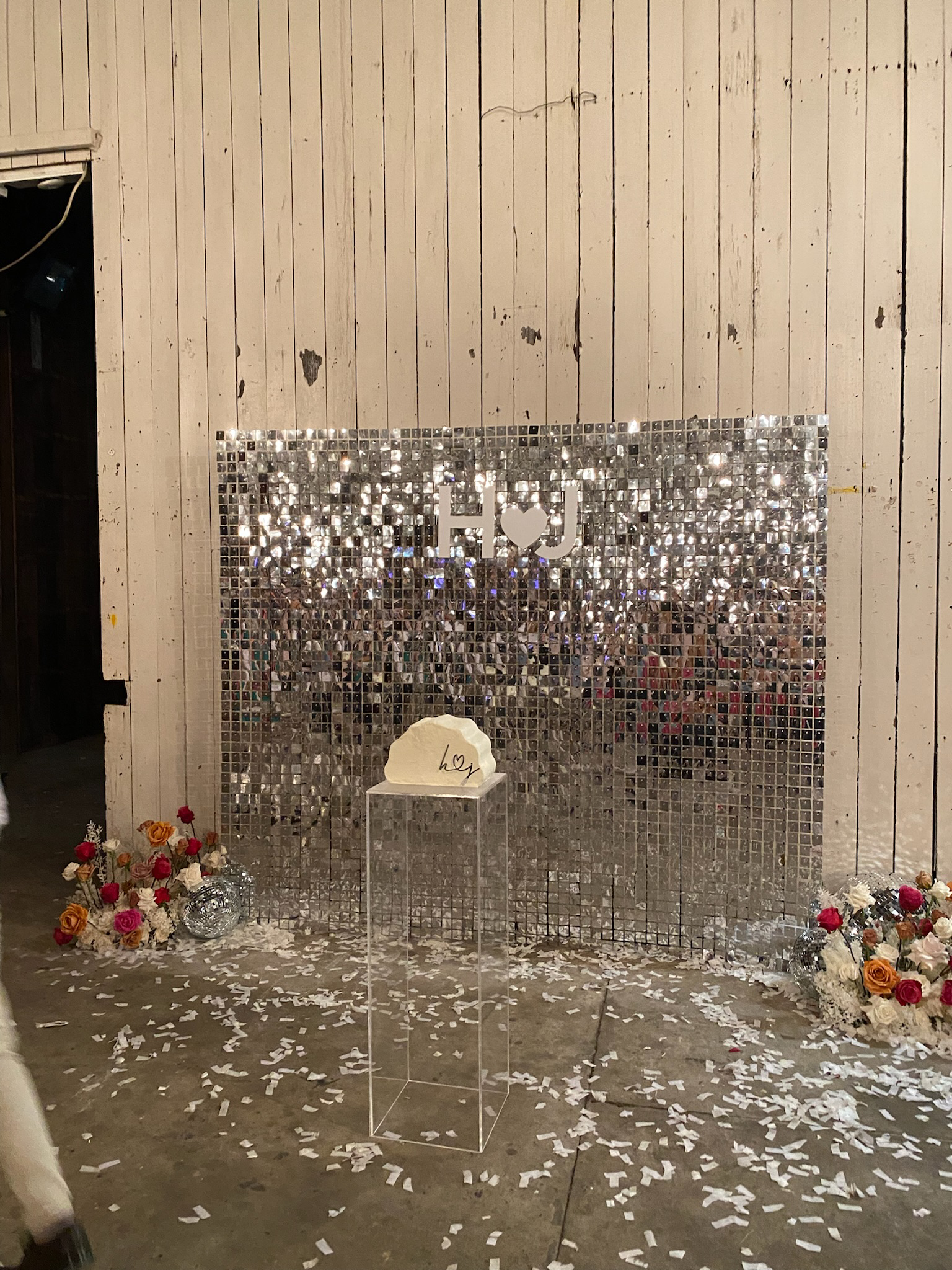 Decorative backdrop with a shiny mirror tile wall, flowers on either side, a transparent stand with a heart-shaped sign, and confetti on the ground for a celebration.