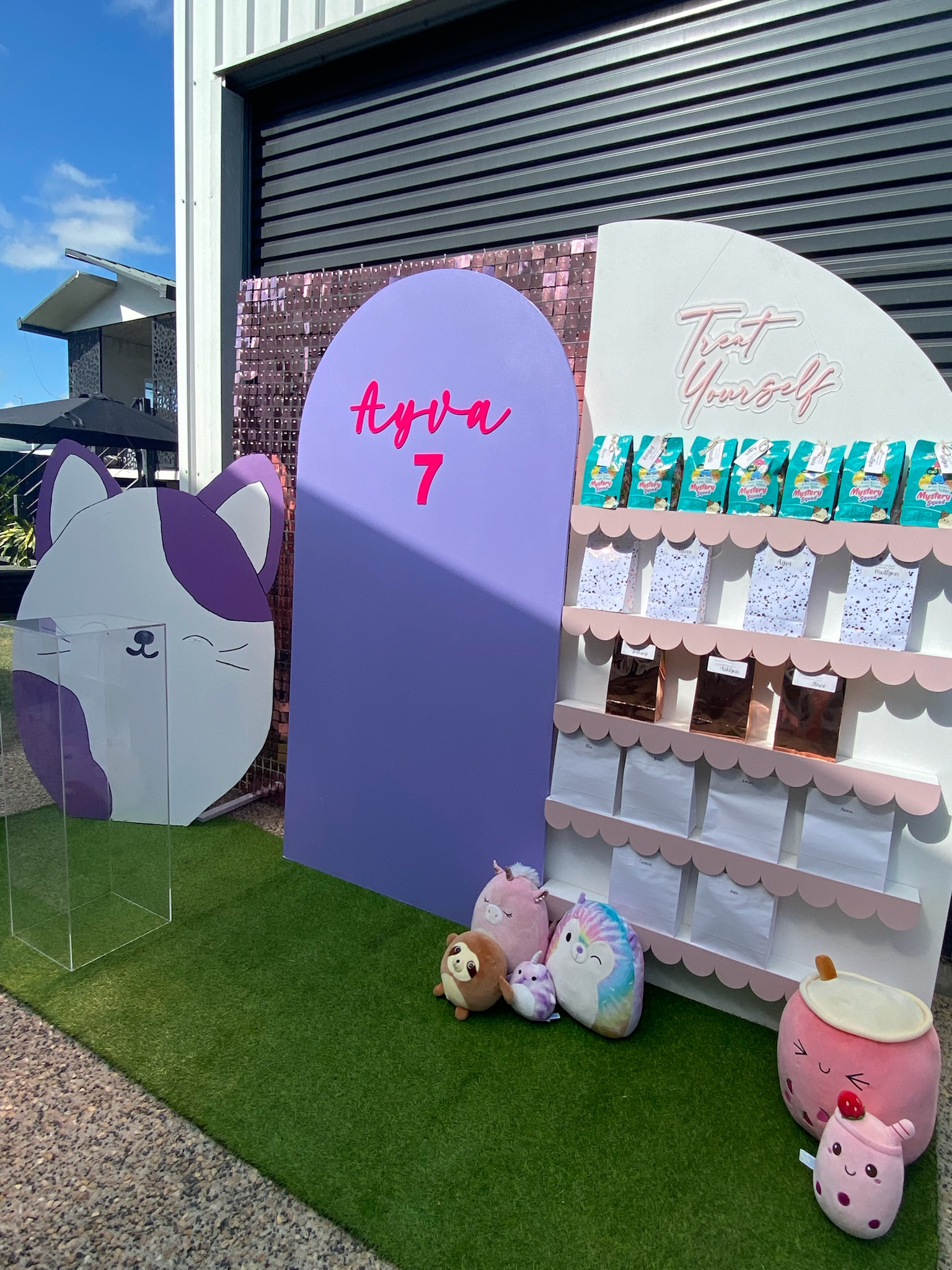 Colorful display with plush toys including a unicorn, sloth, rainbow unicorn, and others, standing beside a purple backdrop with text 'Aya 7', a white shelf with snacks, and a sign saying 'Text Yourself'.