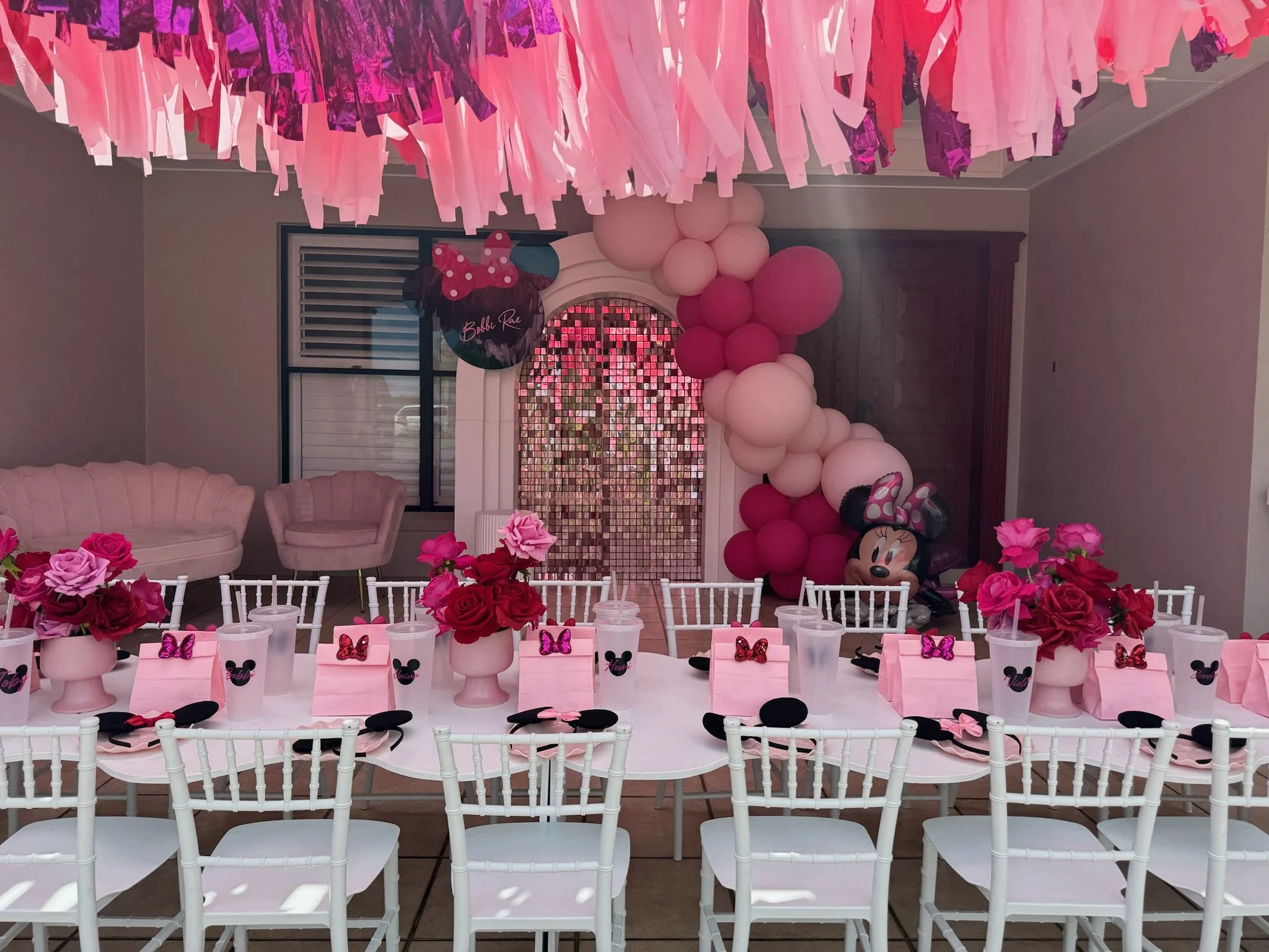 Party room decorated in pink with Minnie Mouse theme, featuring a balloon arch, pink flowers, Minnie Mouse-themed party favors on the table, and Minnie Mouse balloons at the back.