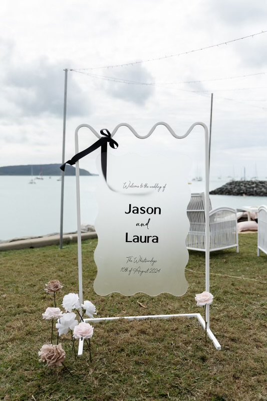 A welcome sign on a stand with flowers at a waterfront, welcoming Jason and Laura on their wedding day, August 14, 2024.