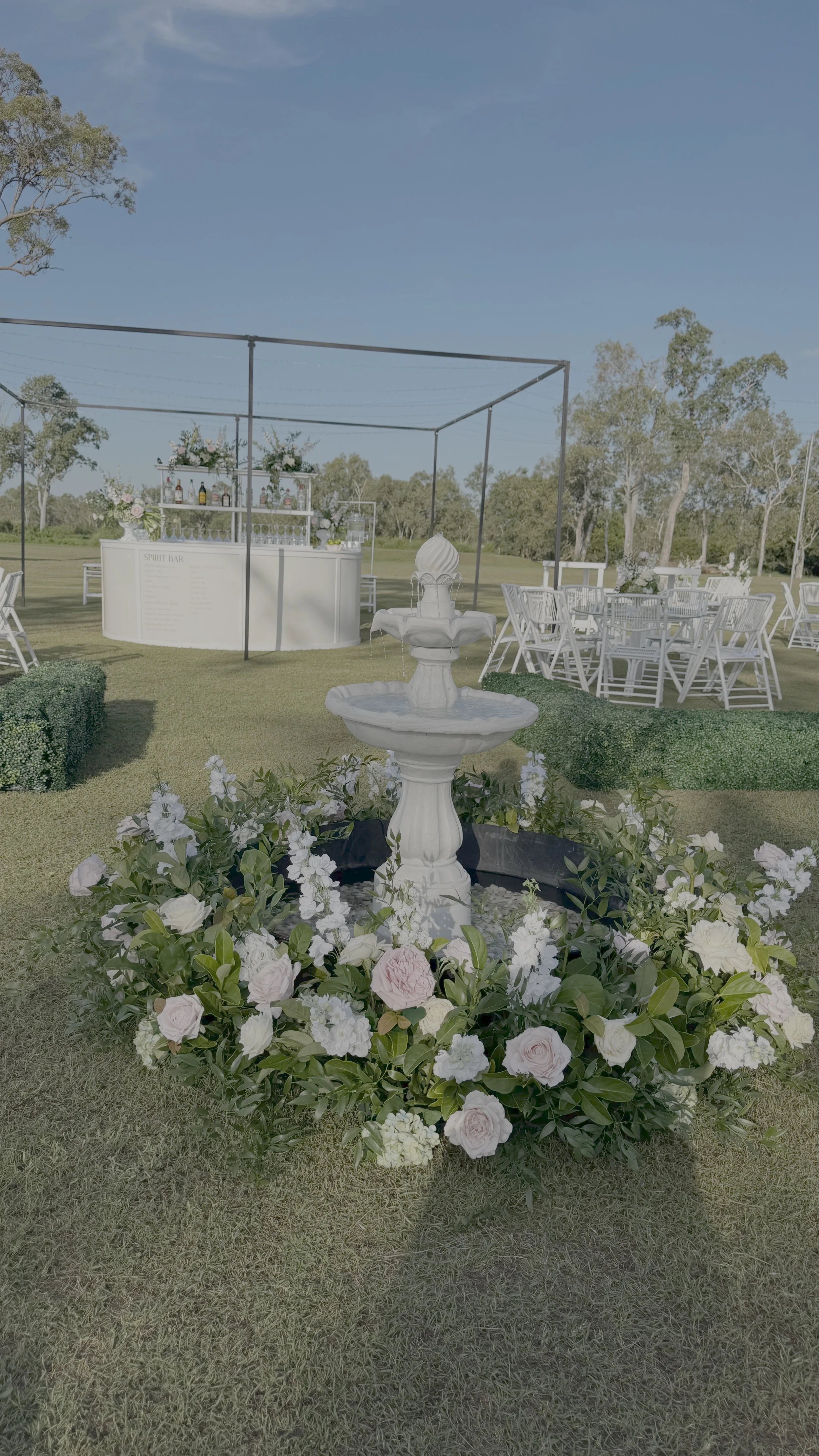 Outdoor wedding or event setup with a white fountain surrounded by white and pink flowers, white chairs, and a bar on a grassy field under a clear blue sky.