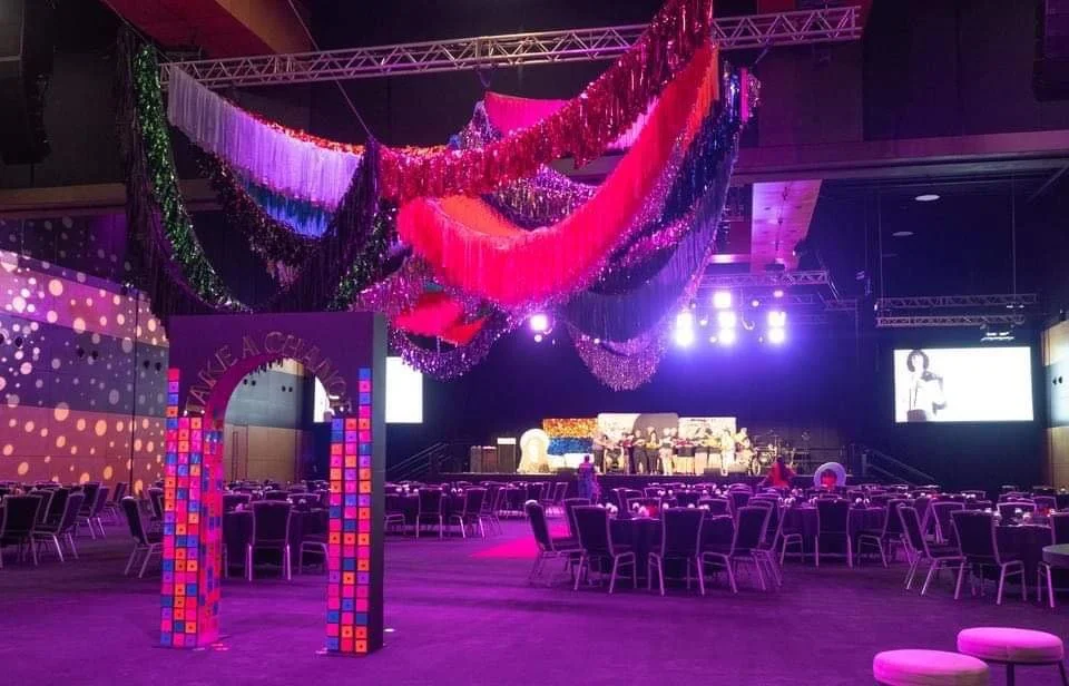 Empty event venue decorated with colorful hanging streamers and purple lighting, with a stage in the background, tables and chairs set up for a celebration.