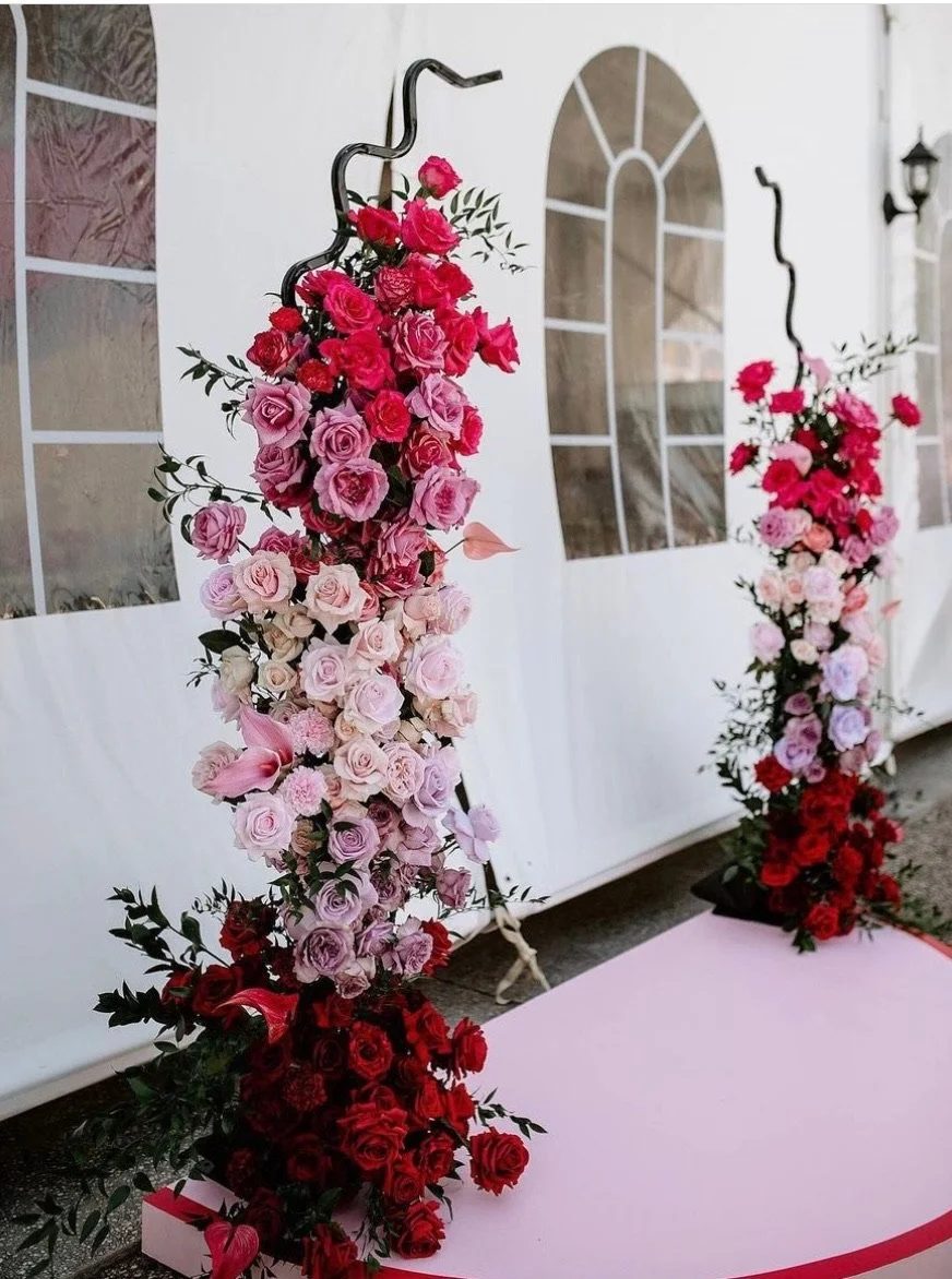 Floral wedding arch decorated with pink and red roses, with black metal stands, set against a white tent with windows and a pink carpeted aisle.