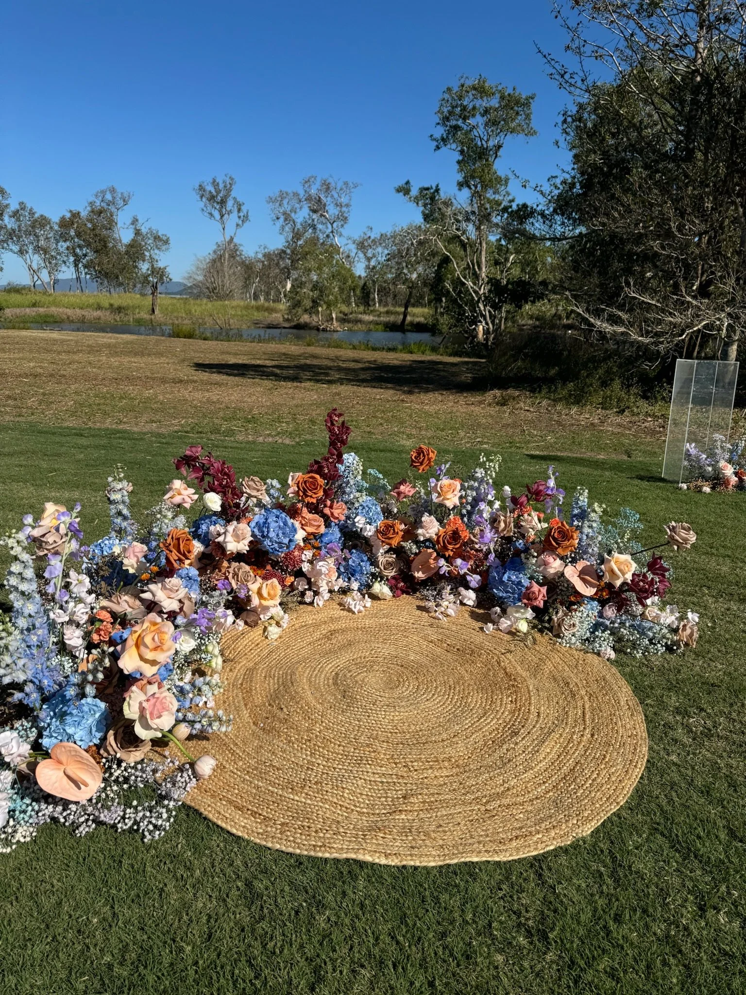 A circular woven rug on grass, decorated with an arrangement of colorful flowers including roses, hydrangeas, and baby's breath, outdoors near trees and a body of water under a blue sky.