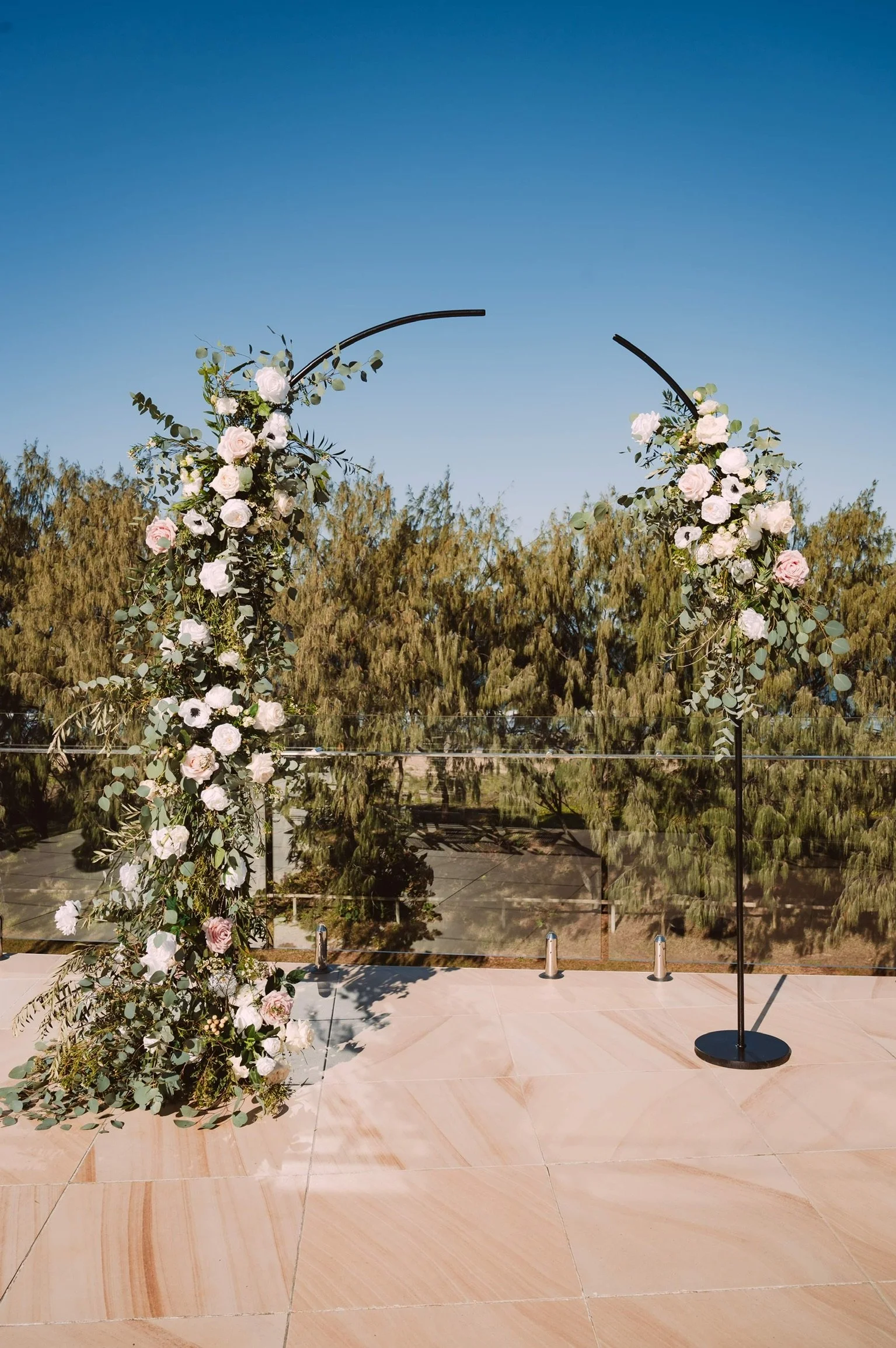A wedding arch decorated with white and blush pink roses, greenery, and eucalyptus leaves, set outdoors on a tiled surface with trees in the background under a clear blue sky.