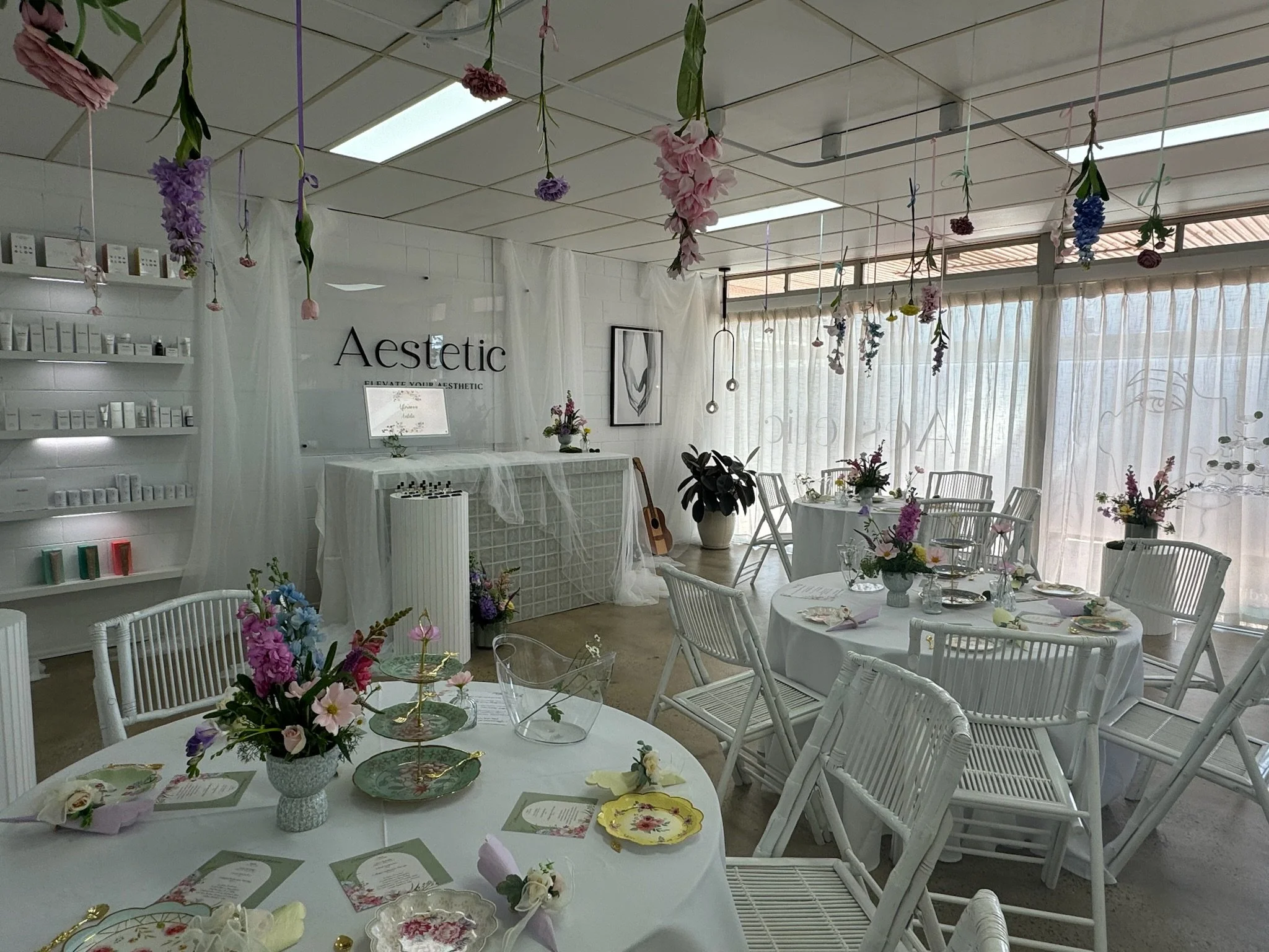 Decorated event space with round tables covered in white tablecloths, floral centerpieces, and place settings. Hanging flowers are suspended from the ceiling, and sheer curtains cover the windows. The space has white chairs, a large potted plant, and