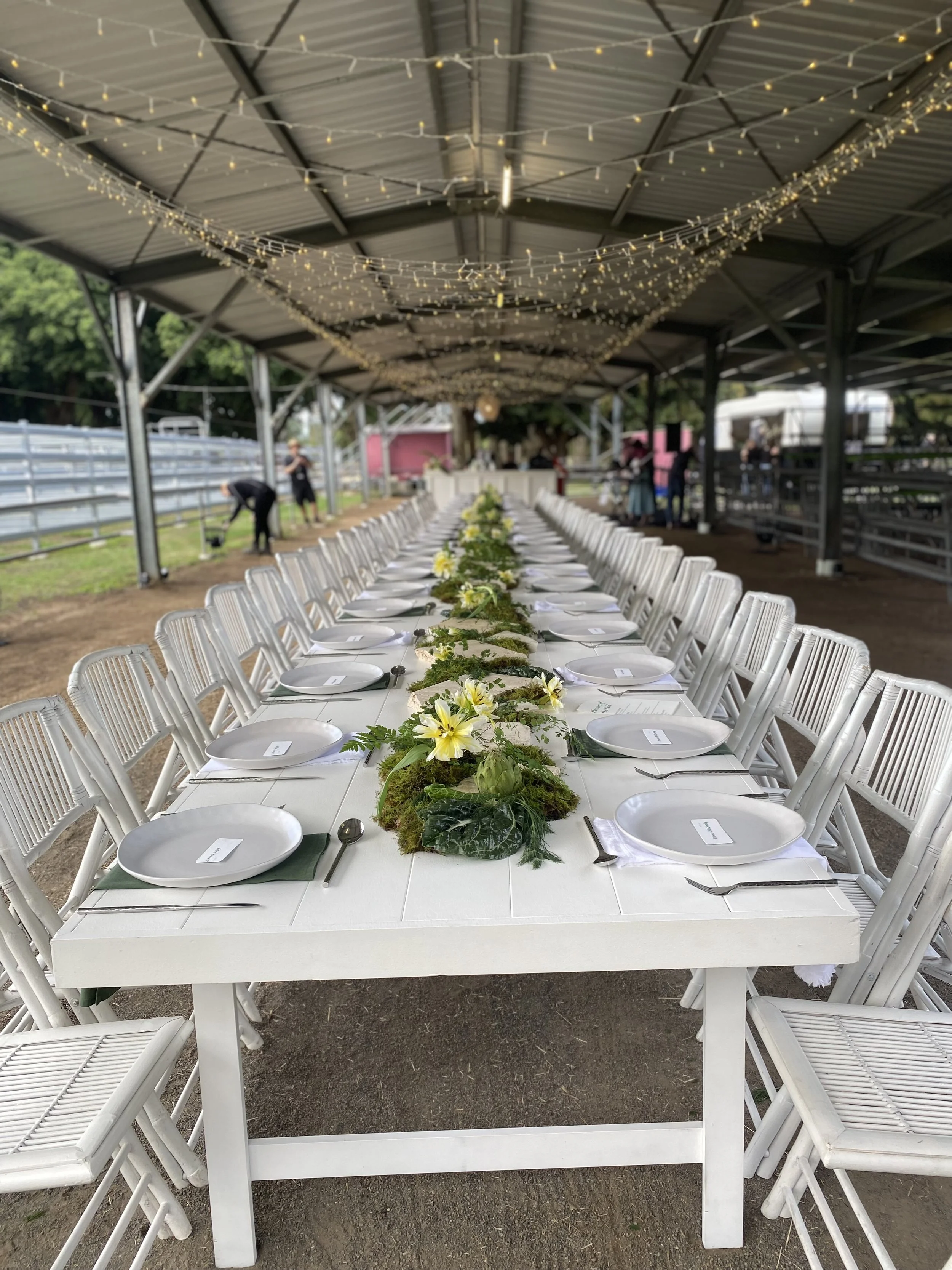 Long banquet table with white plates, silverware, and green napkins, decorated with a green and white floral centerpiece, set under a covered outdoor area with string lights.