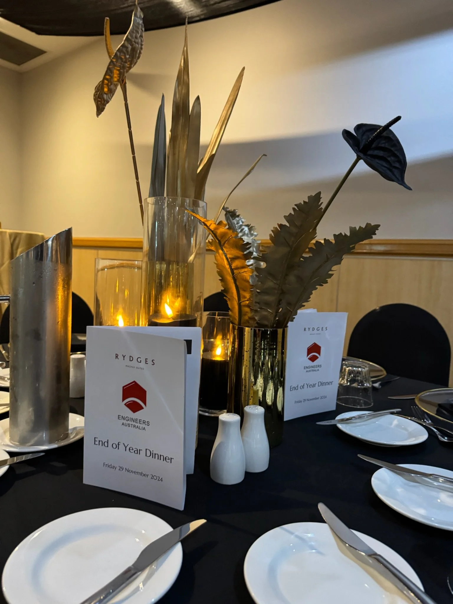 Table centerpiece with gold and black decorative leaves, candles in glass holders, and a small sign reading "End of Year Dinner" at a formal event.