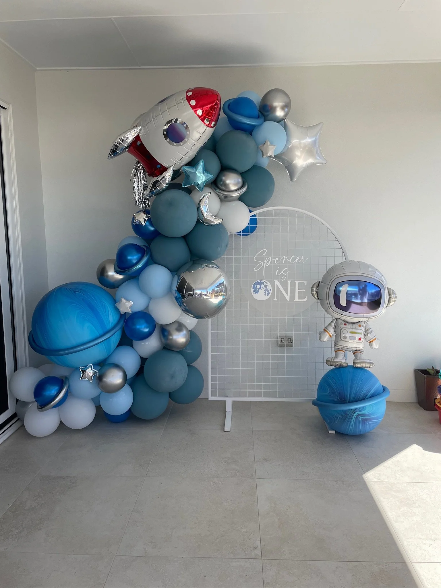 Space-themed birthday decoration with a cluster of blue, silver, and white balloons, a rocket-shaped balloon, and a smiling astronaut figure, with a backdrop that reads "Spencer is ONE."