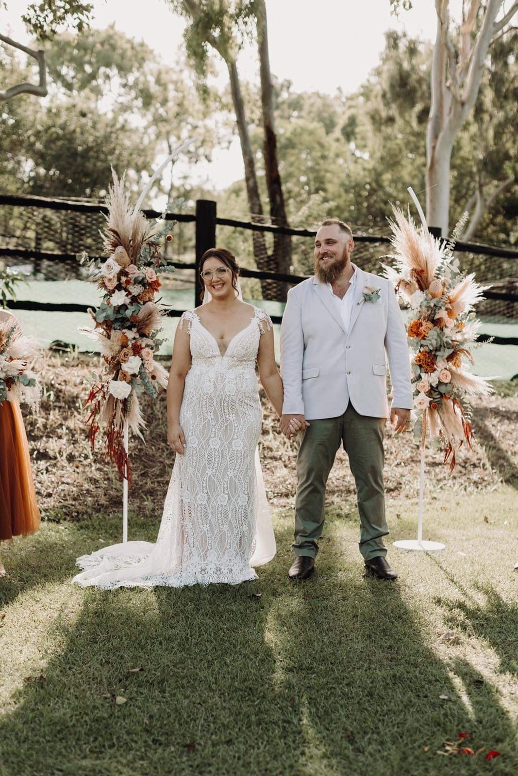 A bride and groom holding hands, standing outdoors among floral decorations with trees and a wooden fence in the background.