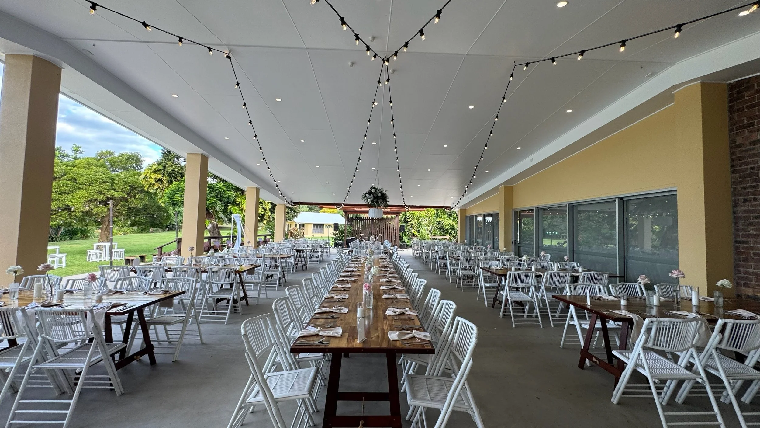 Decorated outdoor banquet area with long wooden tables, white chairs, string lights hanging from ceiling, floral centerpieces, and a view of green trees and lawn outside.