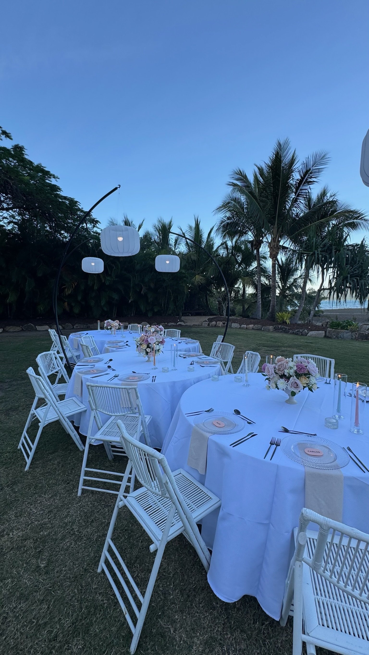 Outdoor banquet setup with white tables and chairs, floral centerpieces, and hanging lanterns, near a tropical landscape with palm trees.