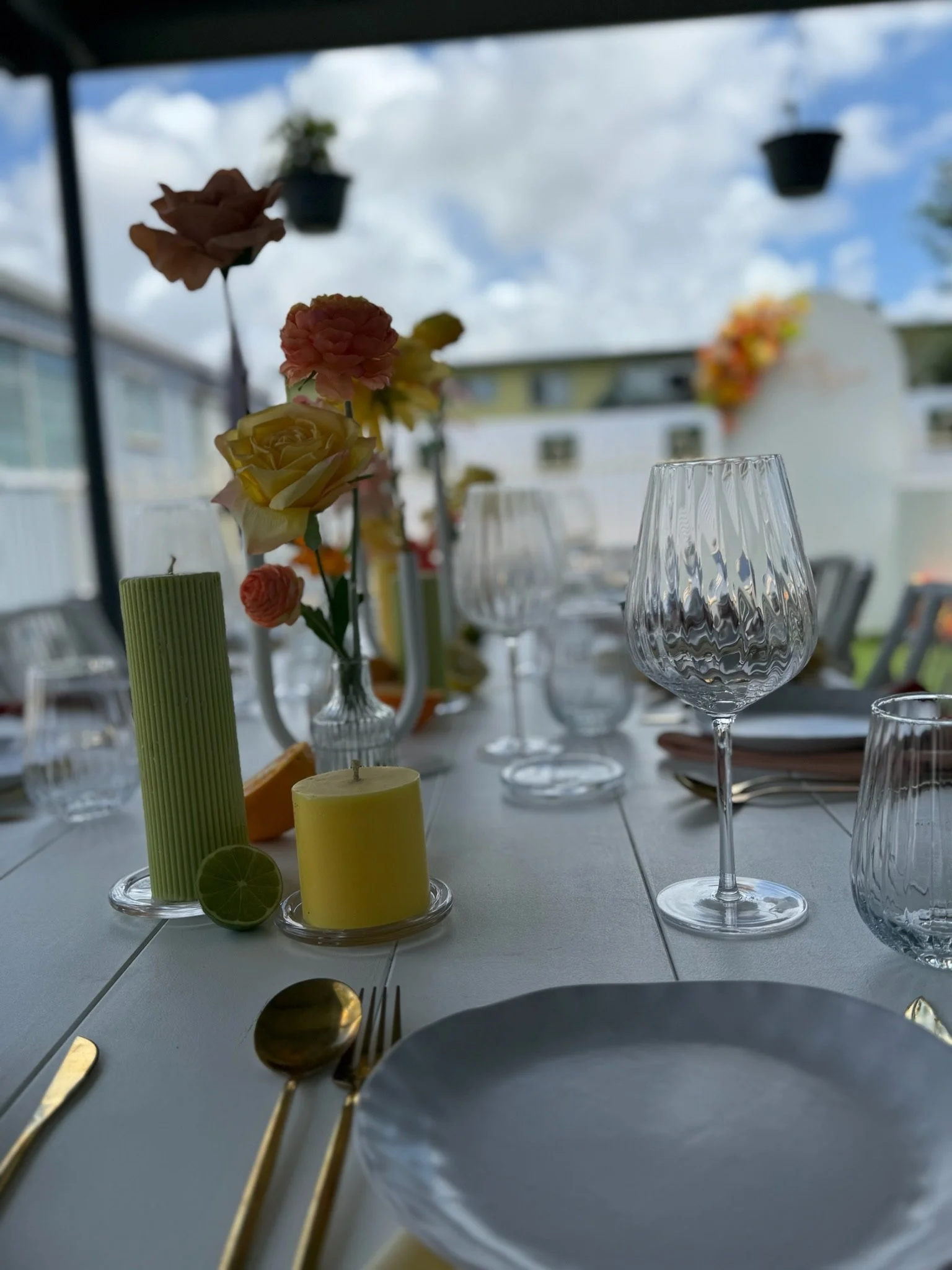 Elegant table setting with colorful flowers, wine glasses, candles, and gold utensils outdoors under a covered patio with a cloudy sky.