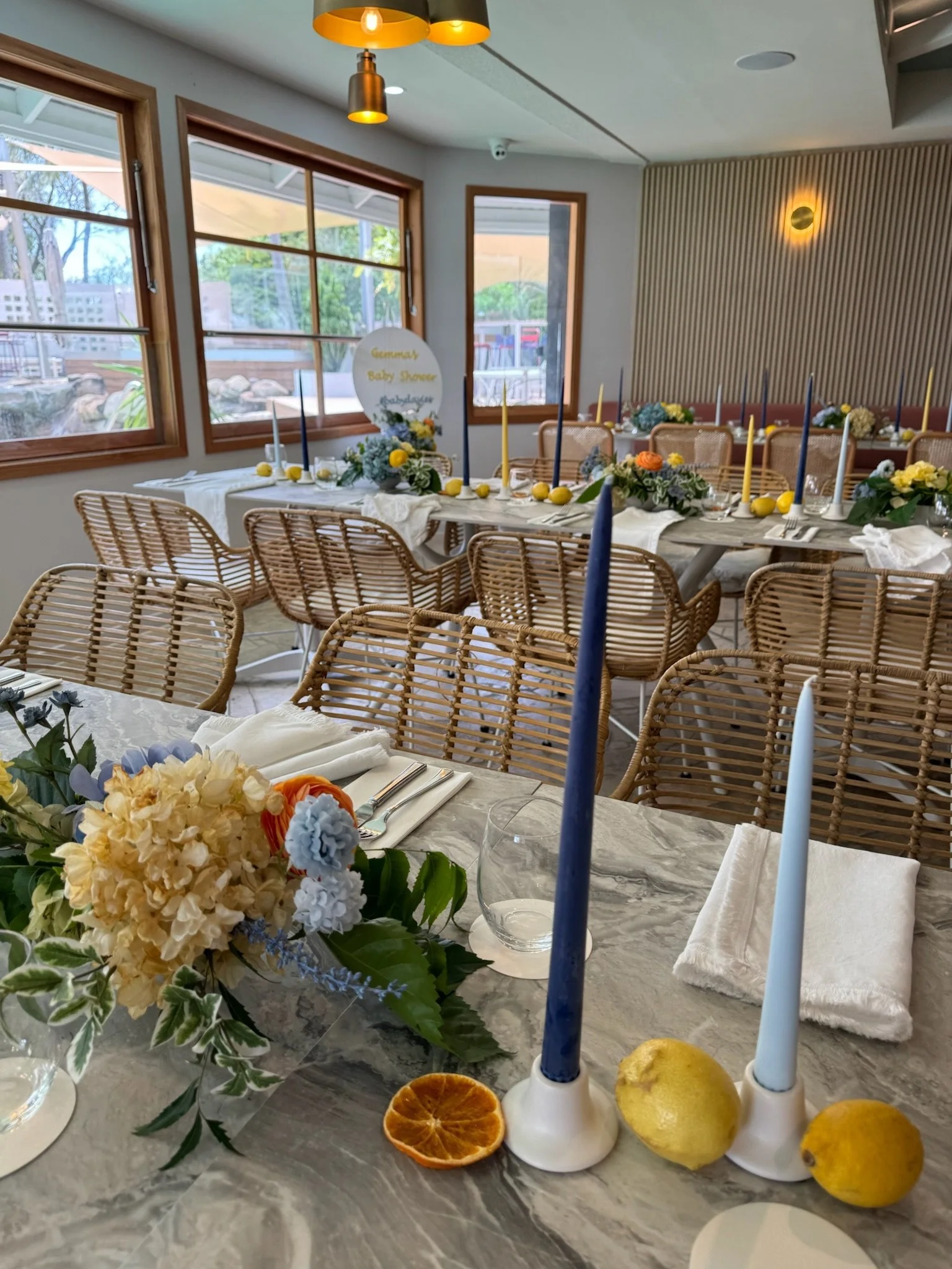 A decorated dining table set for a celebration with floral centerpieces, tall candles, lemons, and a sign that reads 'Summer Baby Shower' in a bright, airy room with windows and wicker chairs.