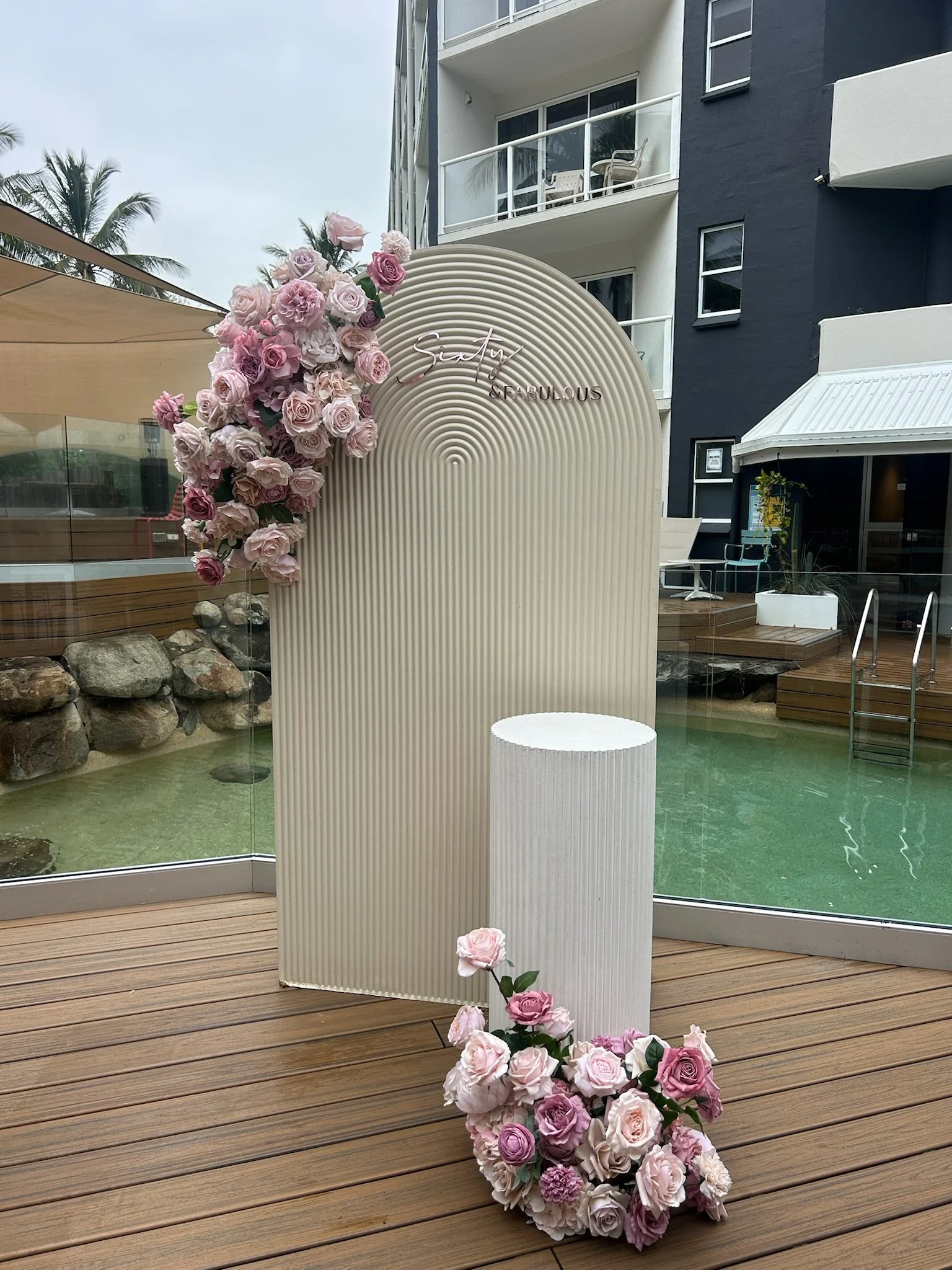 Decorative display with a backdrap of floral arrangements of pink and white roses on a beige curved backdrop with the words 'Sisty & Fabulous' at the top, set on a wooden deck next to a swimming pool in an outdoor setting.