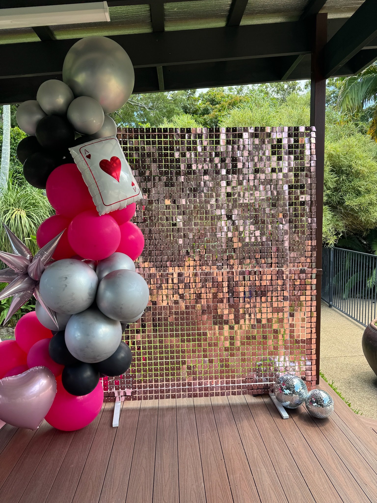 Balloon decoration with pink, black, gray, and silver balloons, including a balloon shaped like a playing card with a red heart, and two disco balls on a wooden stage with a reflective mosaic backdrop surrounded by greenery.