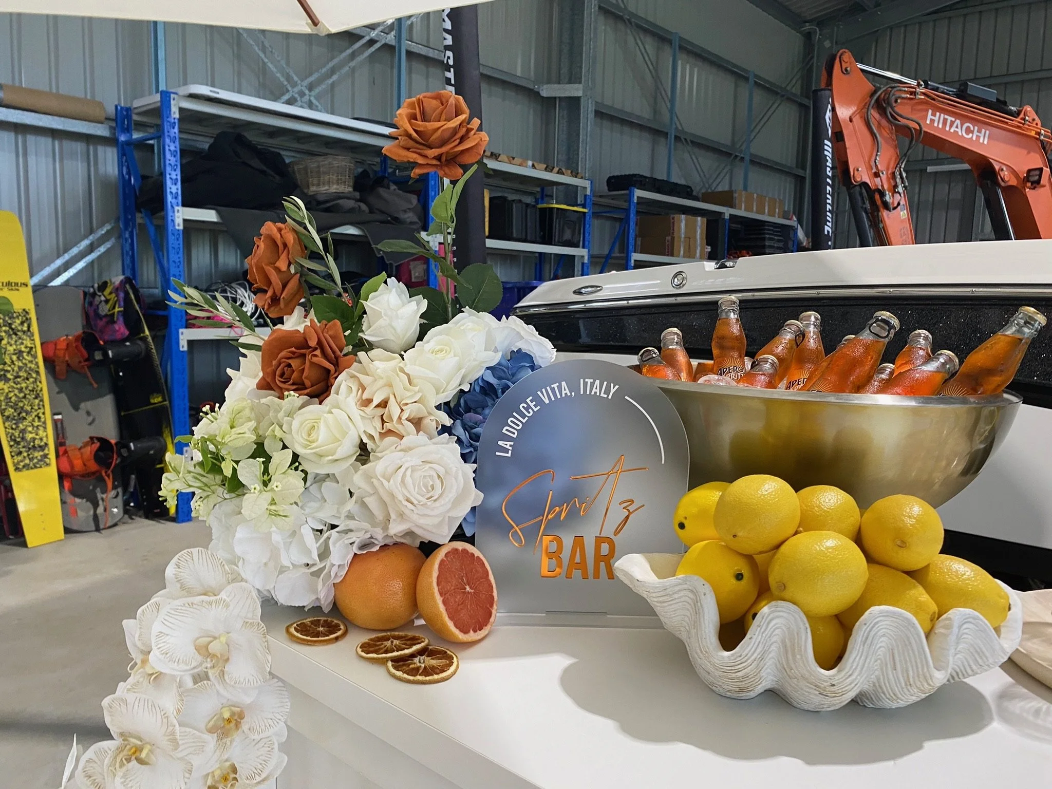 A display of citrus fruits, flowers, and beverages on a table at an indoor event. The citrus includes lemons and grapefruits, some cut open, with a bowl of bottled drinks, and artificial flowers including roses in various colors arranged beside a sig