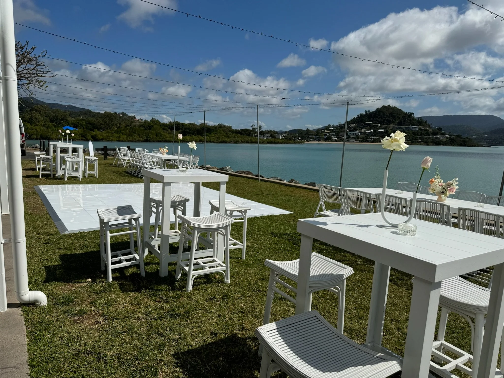 Outdoor event setup with white tables and chairs near a body of water, decorated with vases of flowers, under a partly cloudy sky.