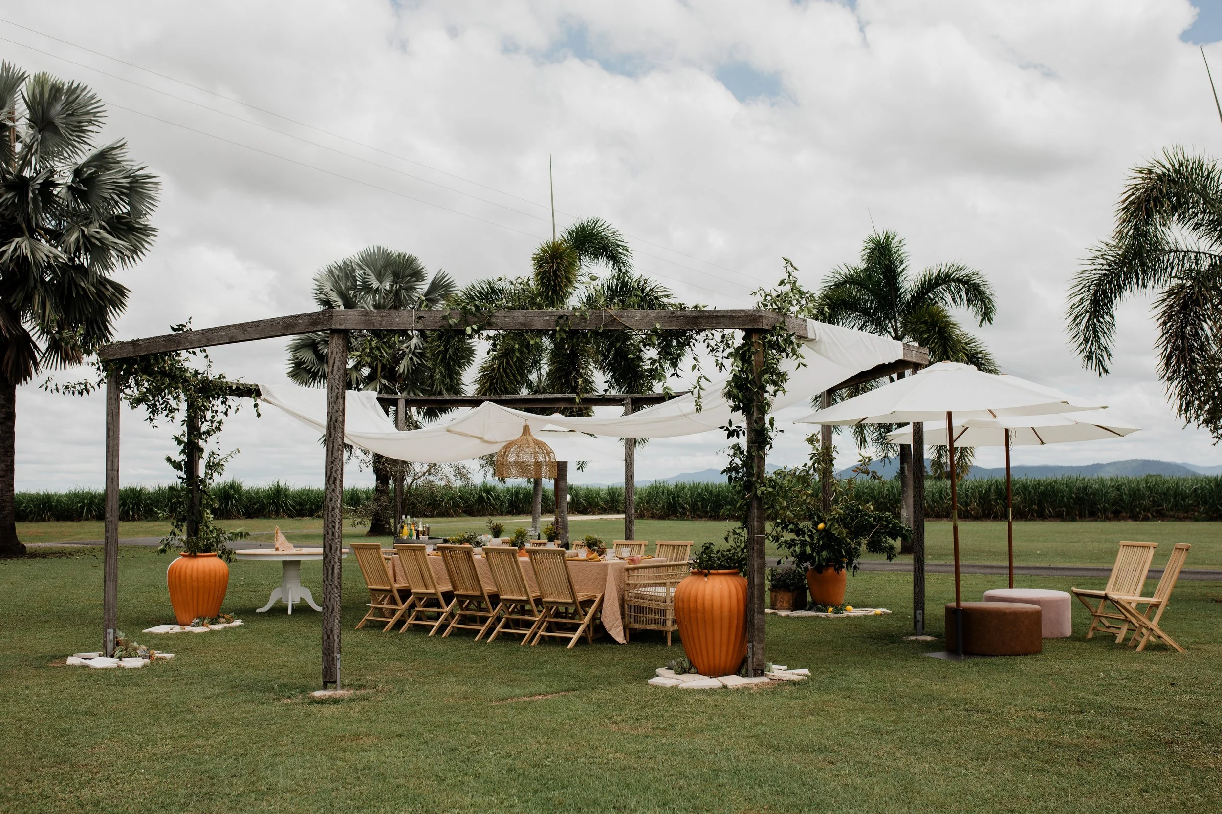 Outdoor dining setup with a long wooden table and chairs under a wooden pergola with draped white fabric, surrounded by large plants and trees, with umbrellas and seating areas on a grassy lawn under cloudy sky.
