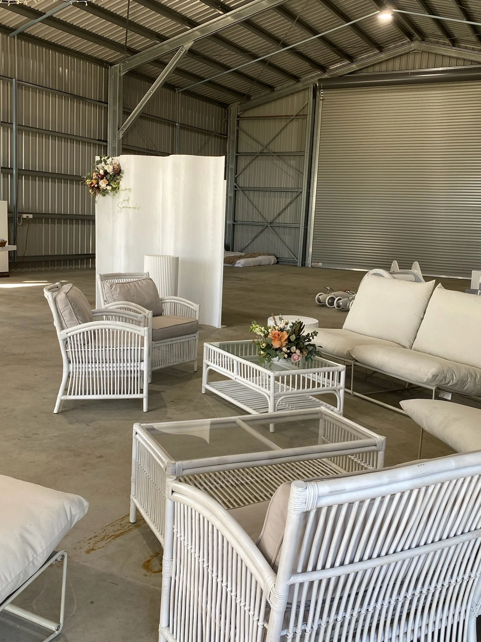 Indoor event space with white wicker furniture, including armchairs, sofas, and tables, decorated with floral arrangements, set up for a wedding or celebration in a large industrial-style warehouse with metal walls and ceiling.