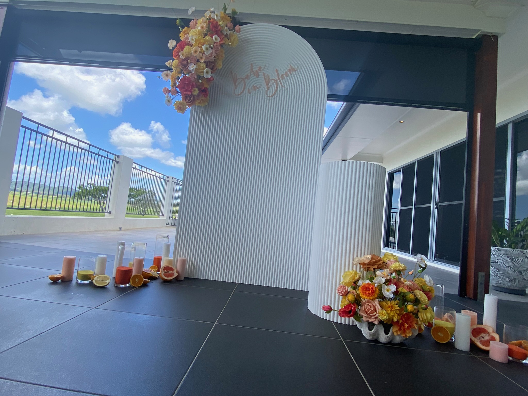 Decorative floral arrangement with candles and citrus slices on a balcony with a view of a blue sky and clouds.