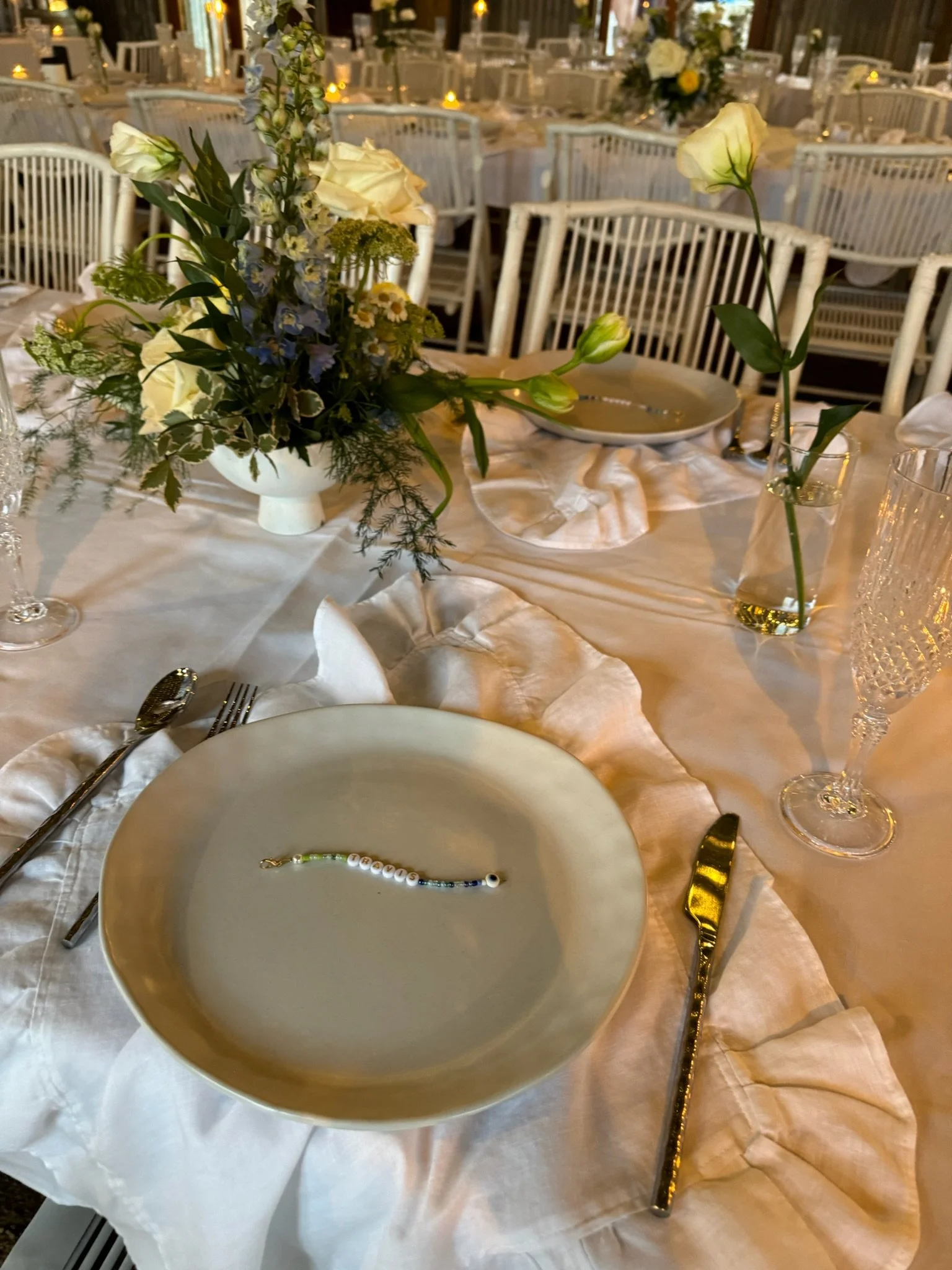 Elegant banquet table set with white tablecloth, floral centerpiece, empty white plate with a beaded bracelet, silverware, and glassware in a decorated event space.