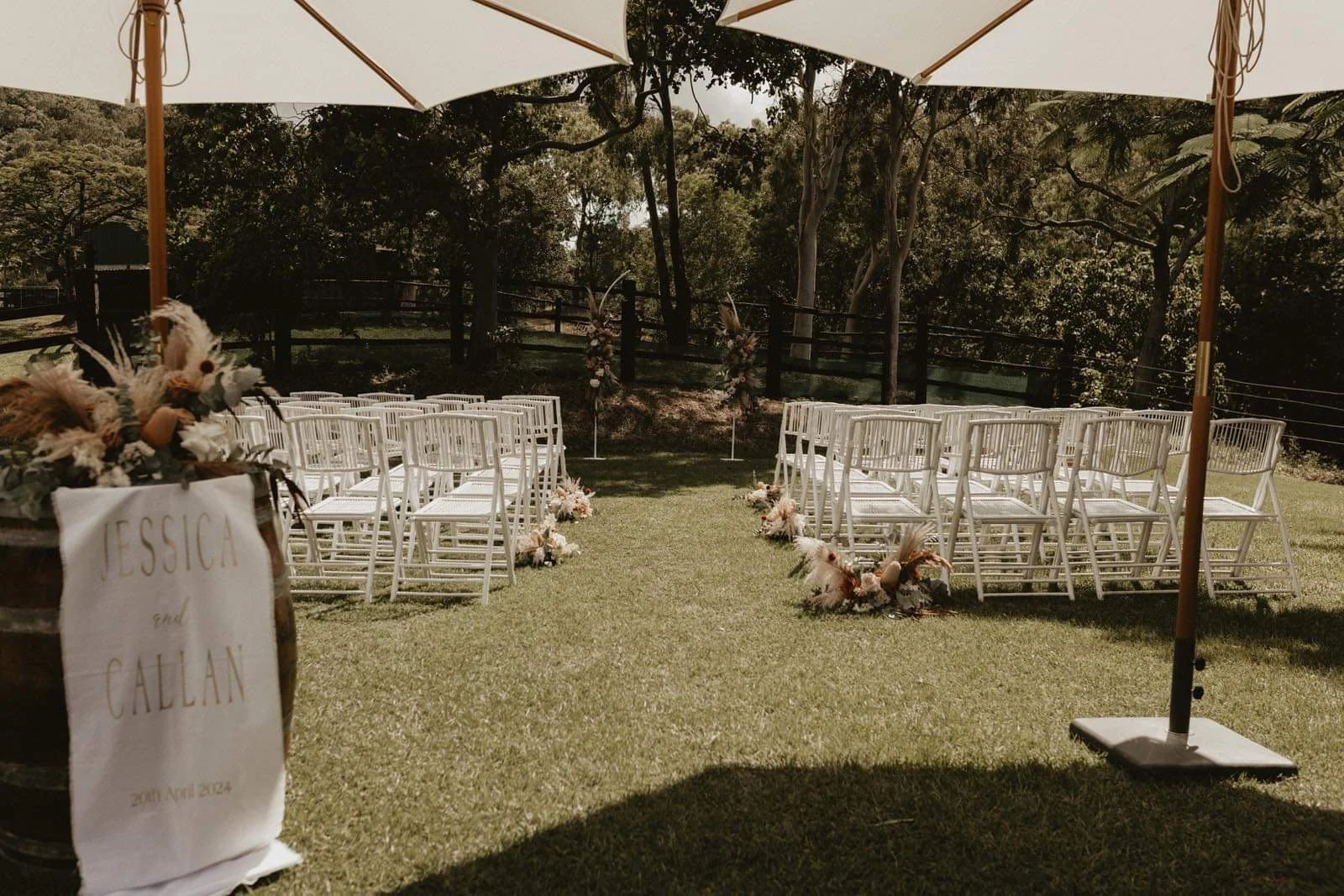 Outdoor wedding ceremony setup with rows of white chairs under large white umbrellas, decorated with floral arrangements, on a grassy area with trees in the background.