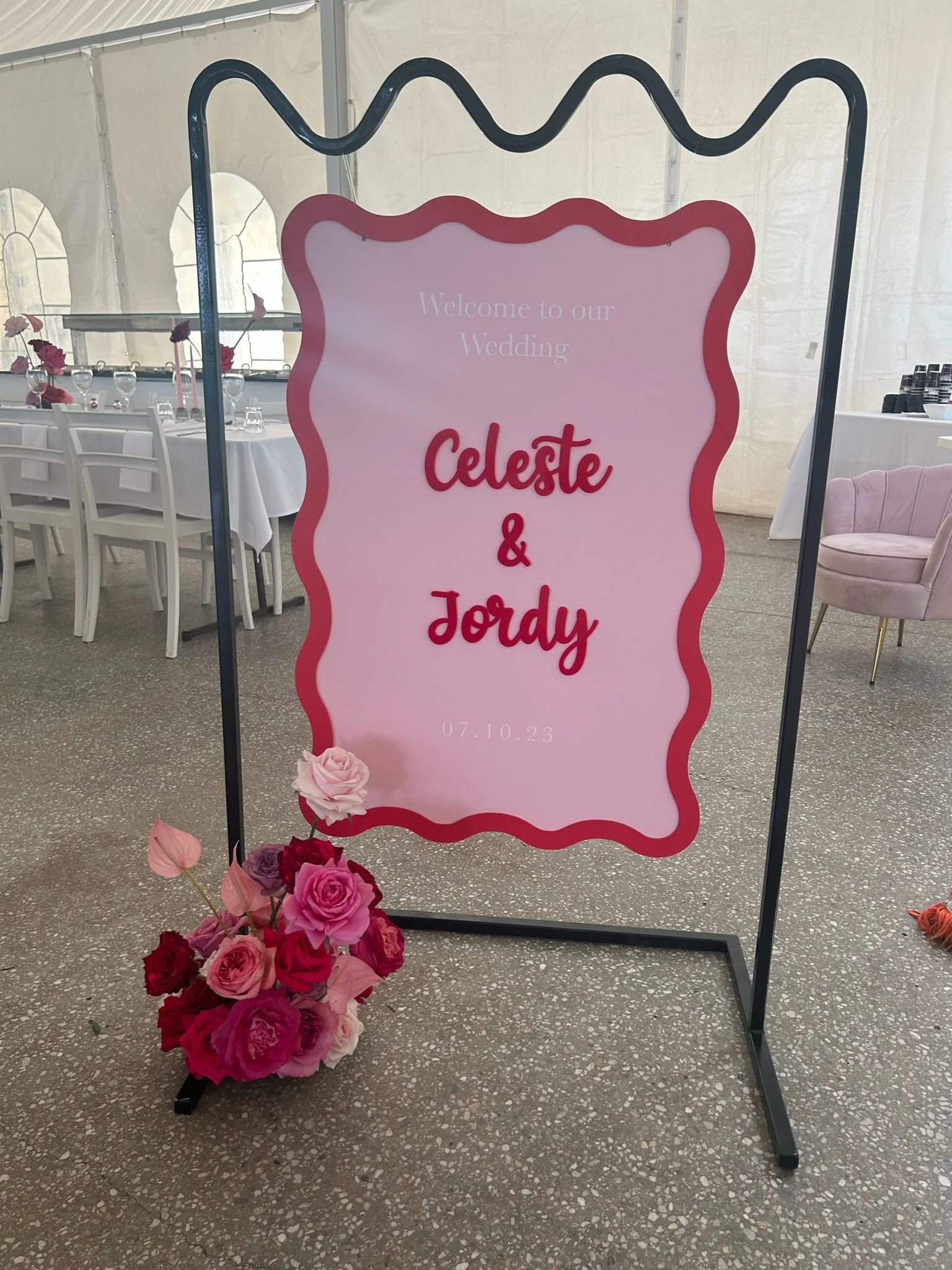 Wedding welcome sign with pink background and red and pink floral decoration, displaying the names Celeste and Jordy, and the date 07.10.23.