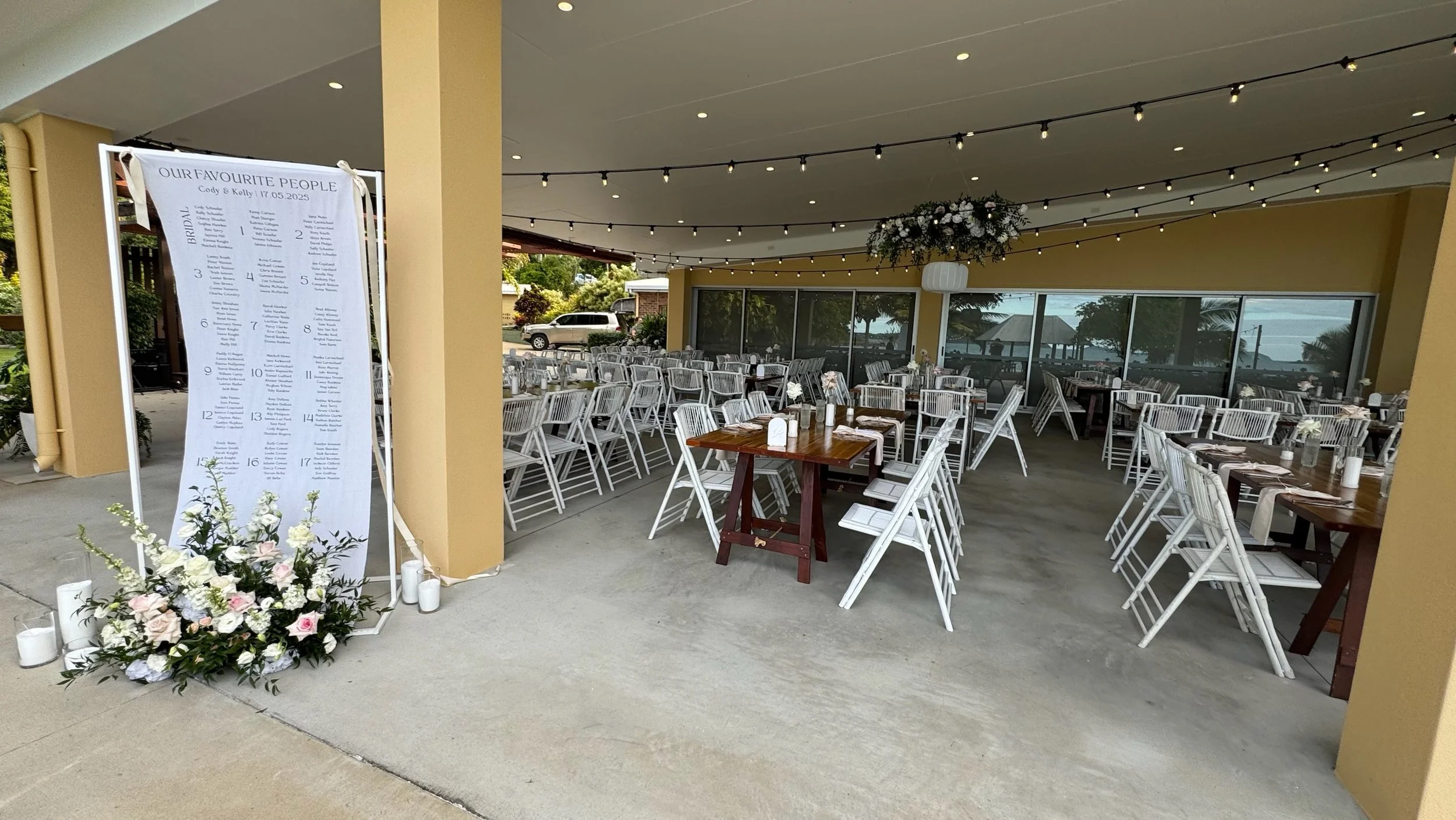 Wedding reception area with arranged tables and white chairs under a decorated covered patio, featuring a large floral centerpiece, string lights, and a seating chart sign.