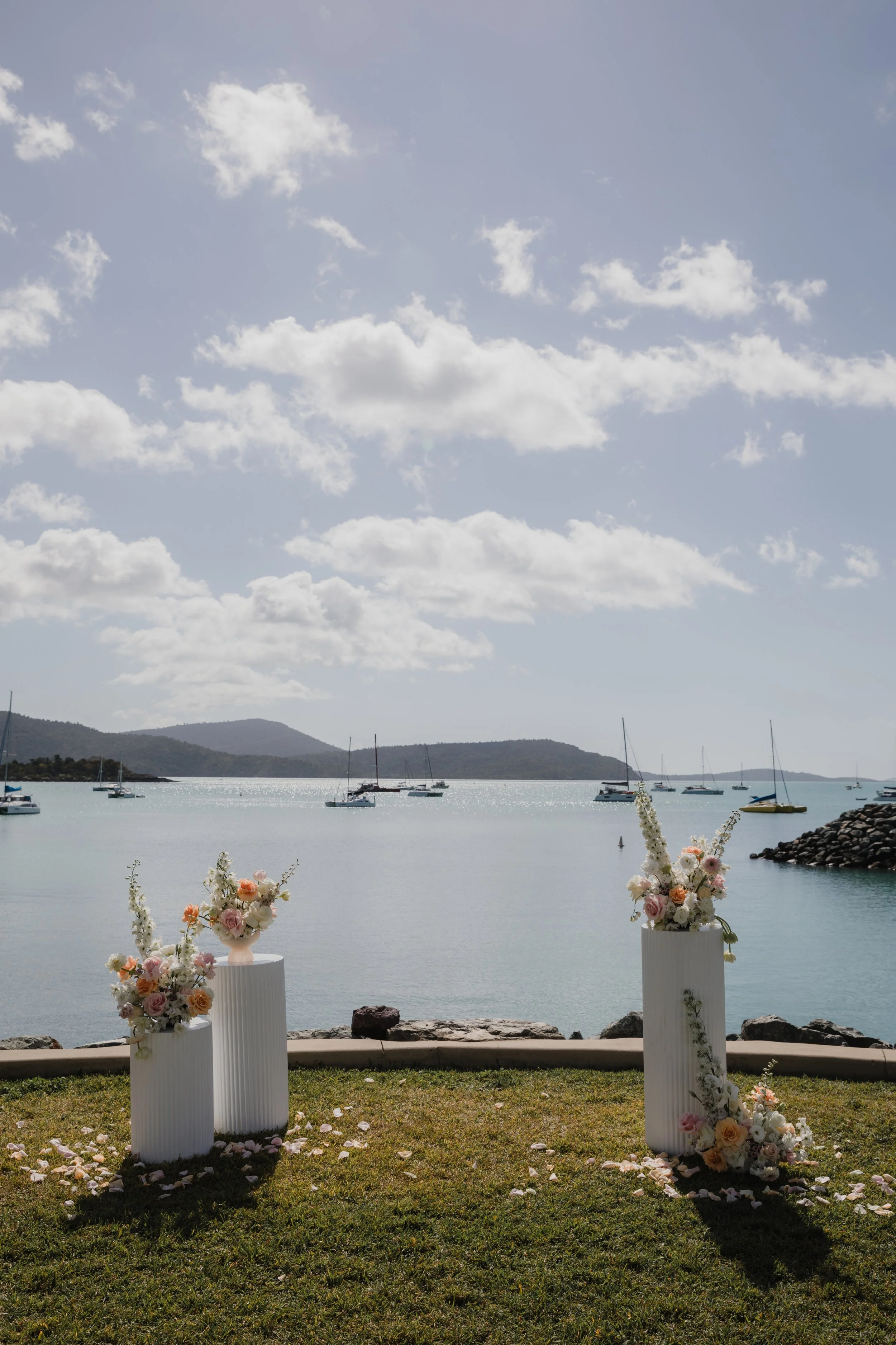 Decorative wedding setup with three white vases filled with pink and white flowers on a grassy area near a body of water, with sailboats on the water and cloudy sky in the background.