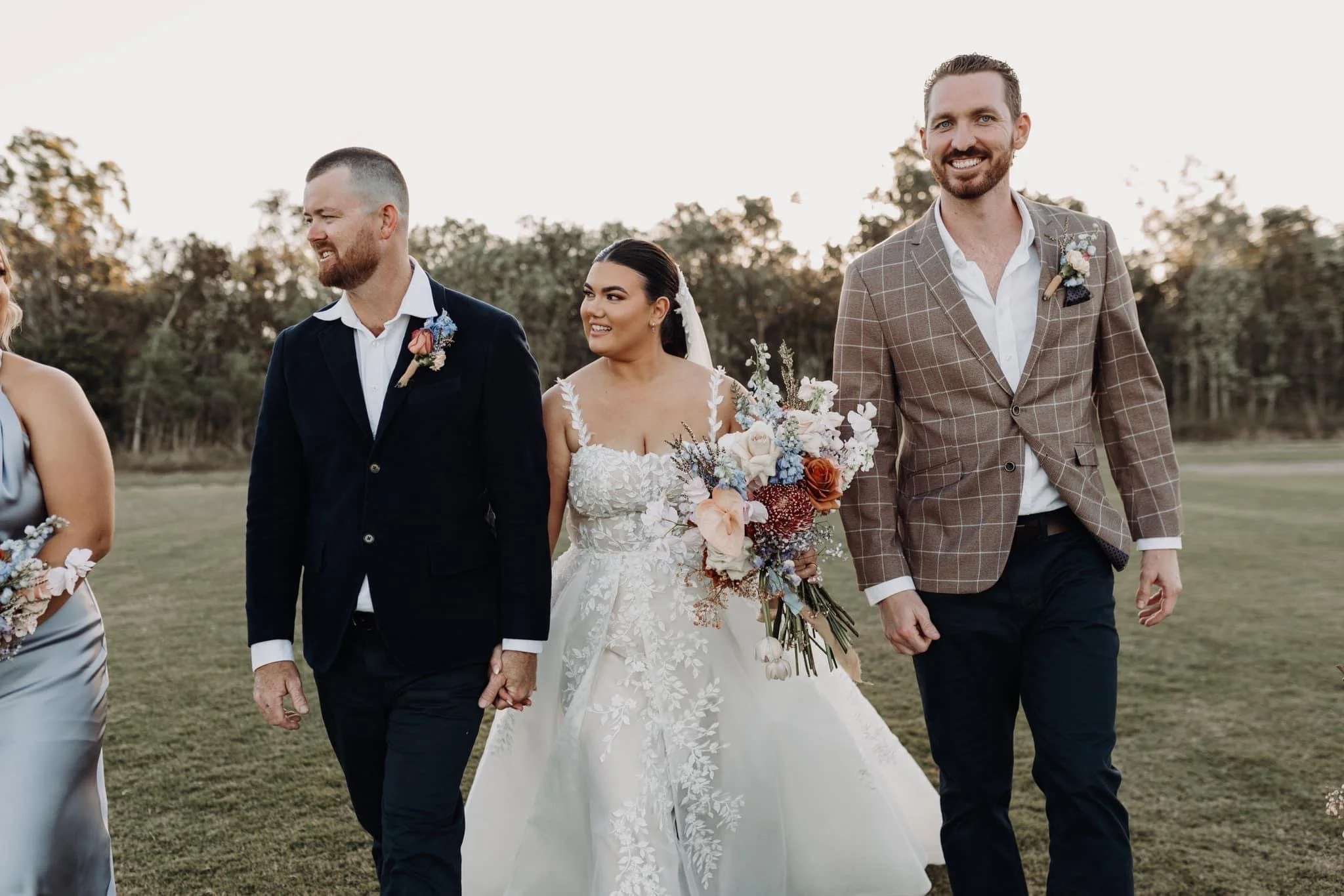A bride in a white wedding gown holding a bouquet of flowers, walking hand-in-hand with two men dressed in suits at an outdoor wedding ceremony during sunset.