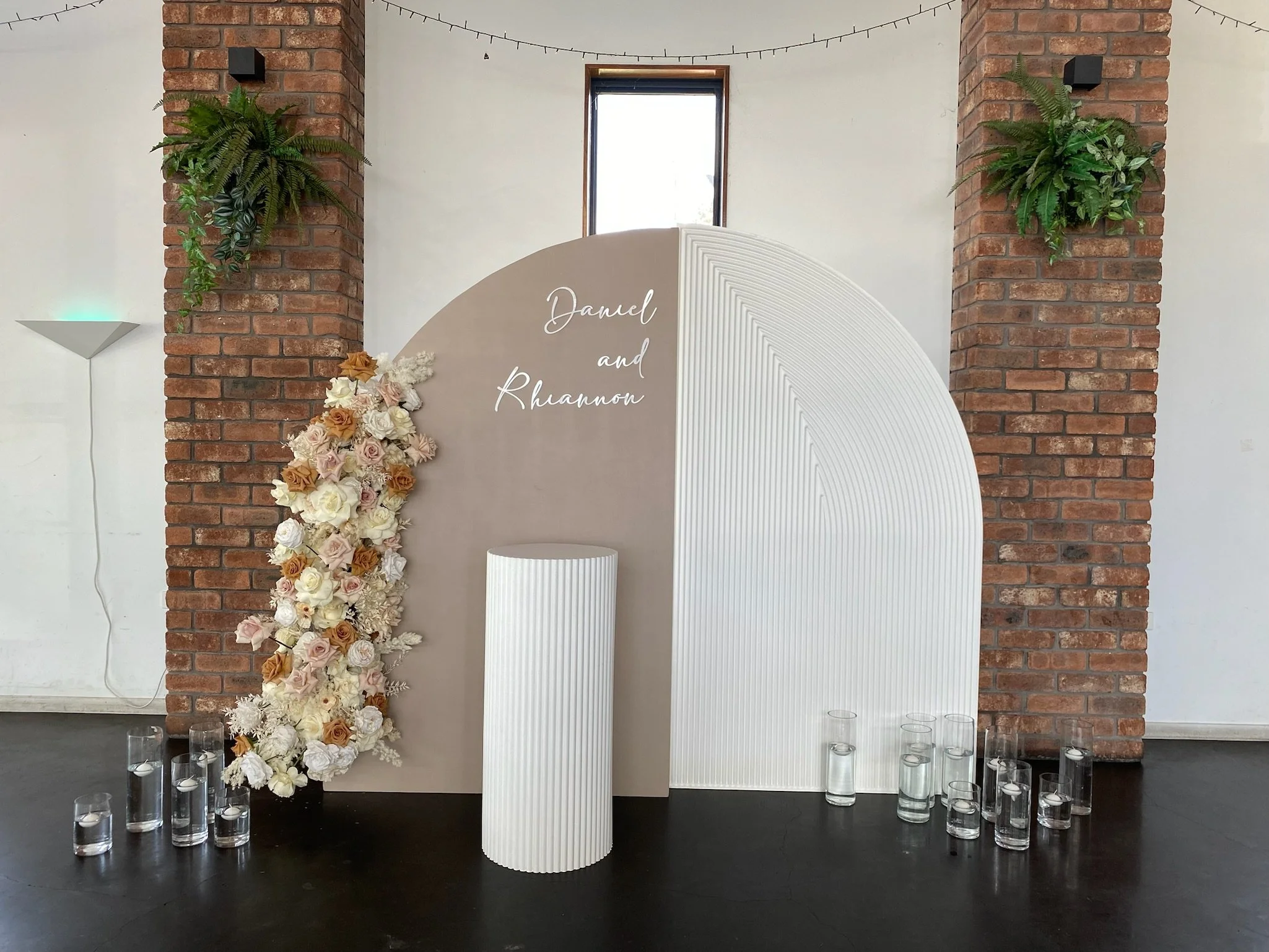 Decorative backdrop with a beige and white panel, floral arrangement on the left, green plants on brick columns, glass vases with water and floating candles at the base, and the words 'Daniel and Rhuannon' written in white script.