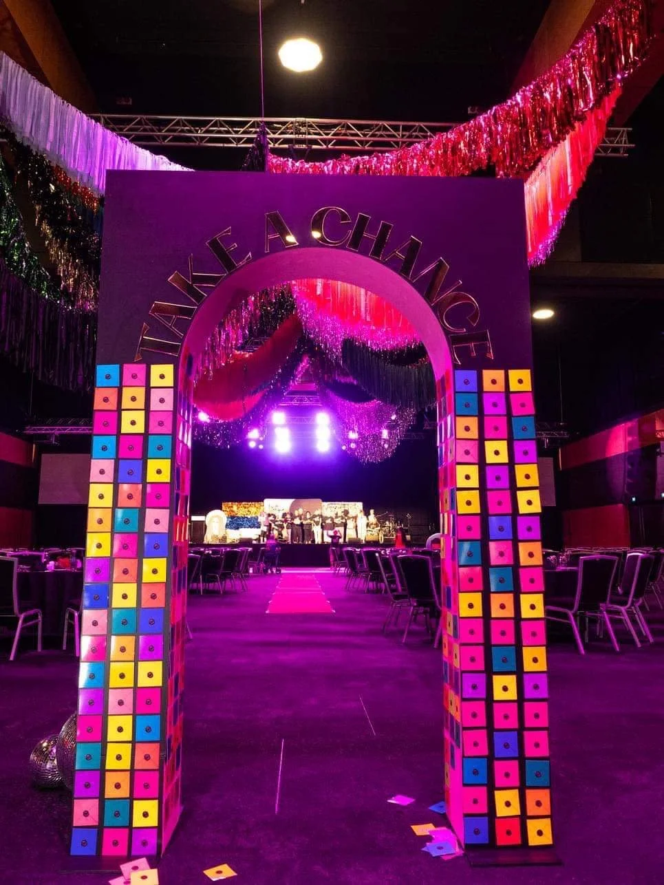 Colorful event stage with an arch decorated with multicolored boxes, leading into a lit stage with performers and large purple lighting, under pink and purple hanging decorations.
