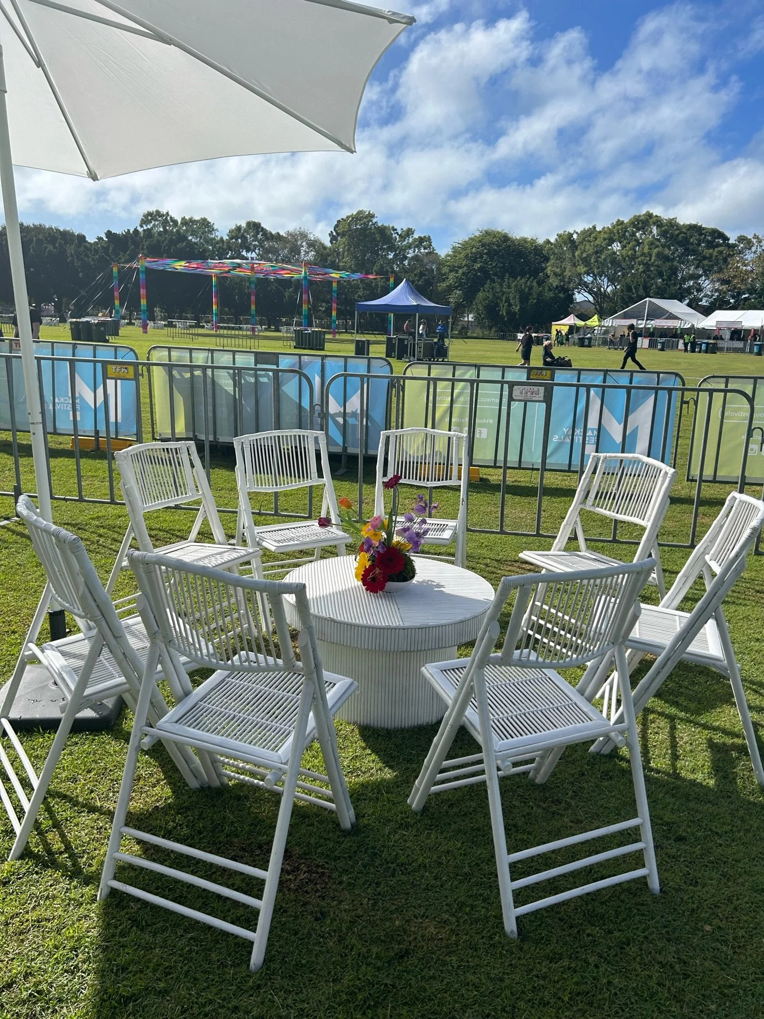 A white round table with a colorful flower arrangement, surrounded by several white chairs, set on green grass at an outdoor event under a large white umbrella. In the background, there are tents, festival booths, and a ferris wheel at a fair or carn