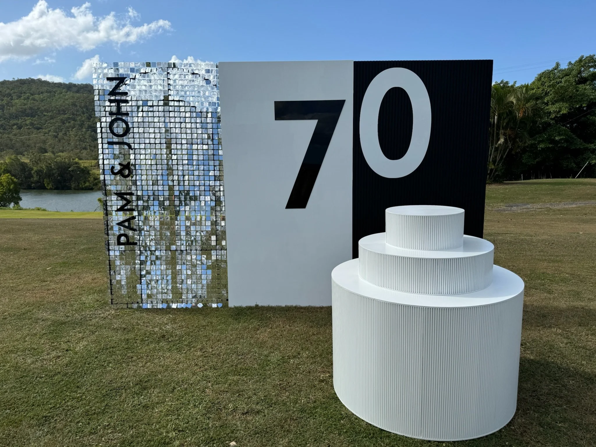 A large outdoor sign celebrating the 70th anniversary of Pam & Joe, with the number 70 in bold black and white, and modern white tiered display stands in front of it, set against a grassy area with trees and a lake in the background.