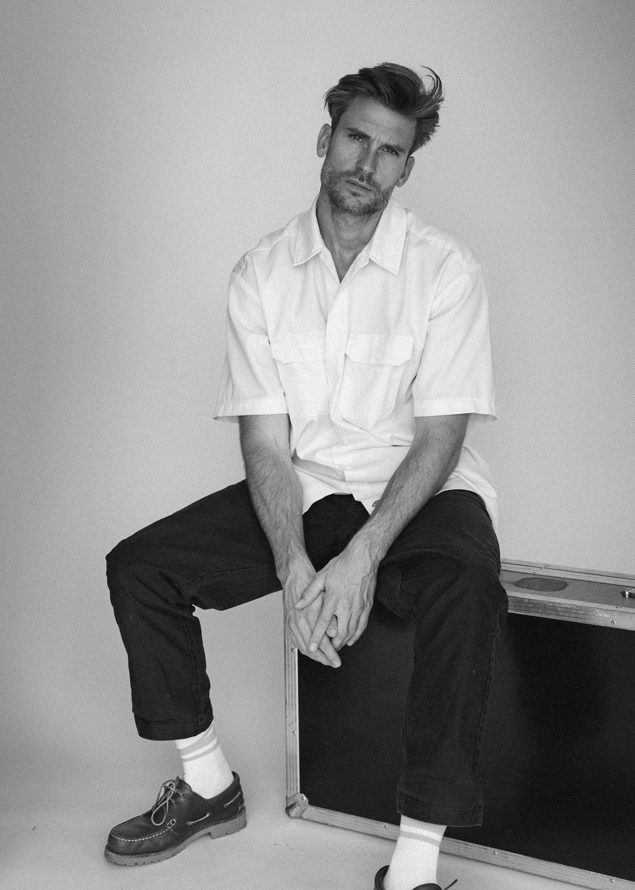 A black and white photo of a man sitting on a box or case against a plain background. He has messy hair, a beard, and is wearing a short-sleeved, button-up shirt, black pants, white socks, and black shoes. He has a serious or contemplative expression on his face.