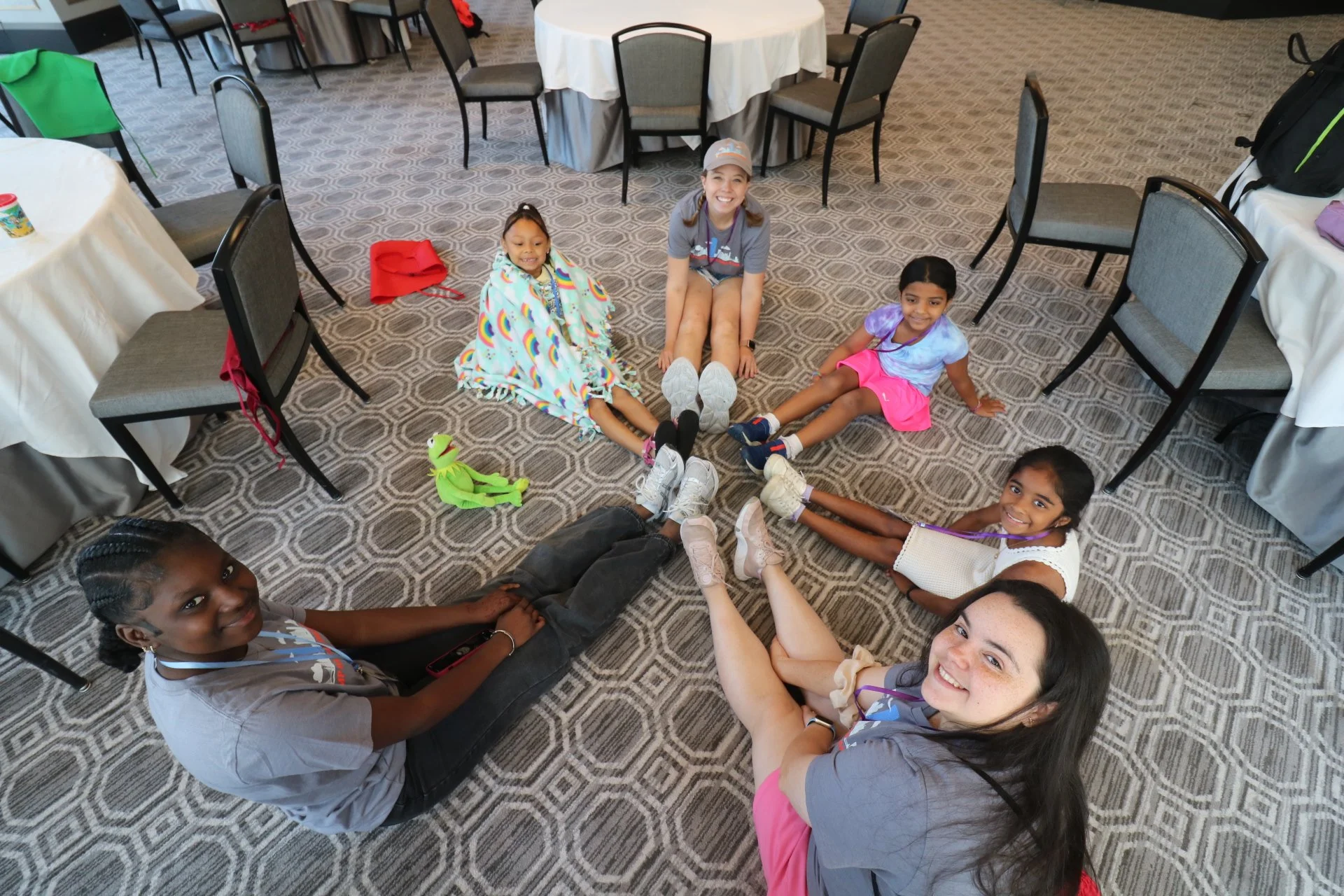 Six people, five children and one adult, sitting on the carpet in a circle with their legs extended and touching each other's feet in a connecting pose, inside a room with round tables and chairs. The children are smiling, and the adult is also smiling.