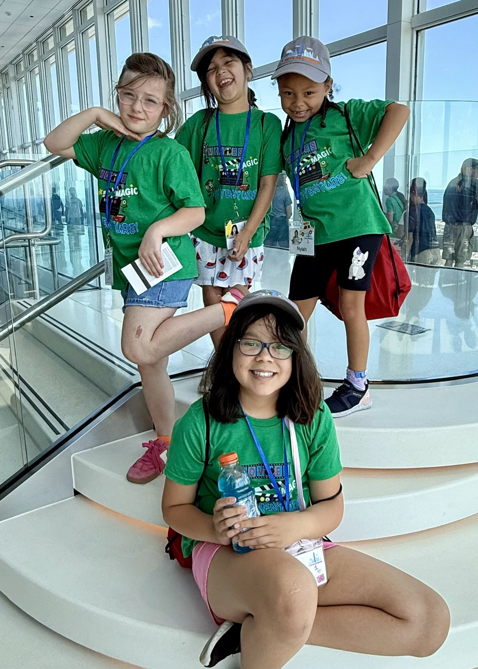 Four children wearing green camp T-shirts with a travel-themed design, posing in an indoor setting with large windows and a glass wall, some wearing hats, badges, and holding water bottles, smiling and making playful poses.